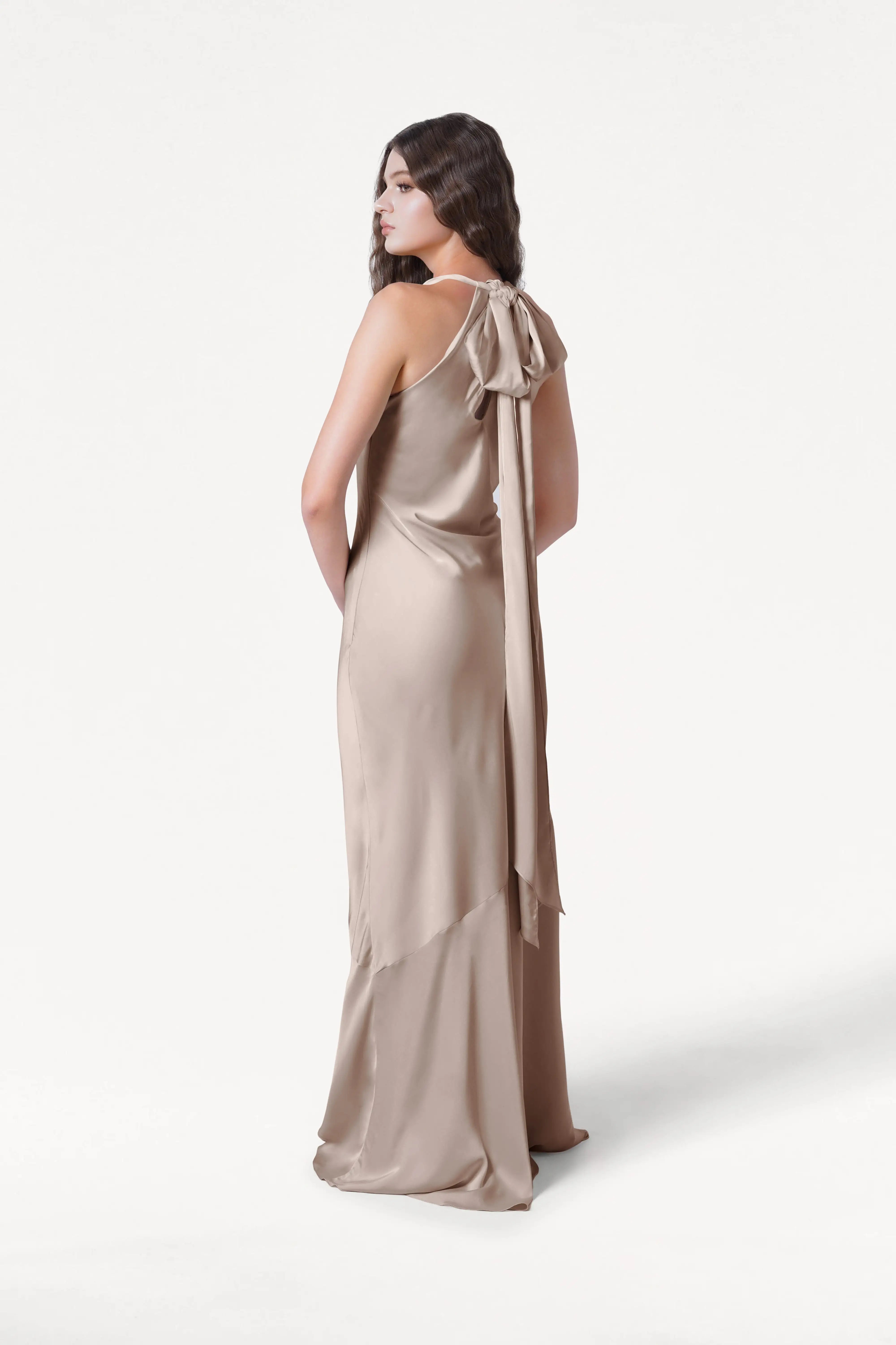 A woman models the TH&TH Mira Bridesmaid Dress in Champagne Sky Satin, featuring a bow at the back, as she stands facing away and looks over her shoulder against a plain white background.
