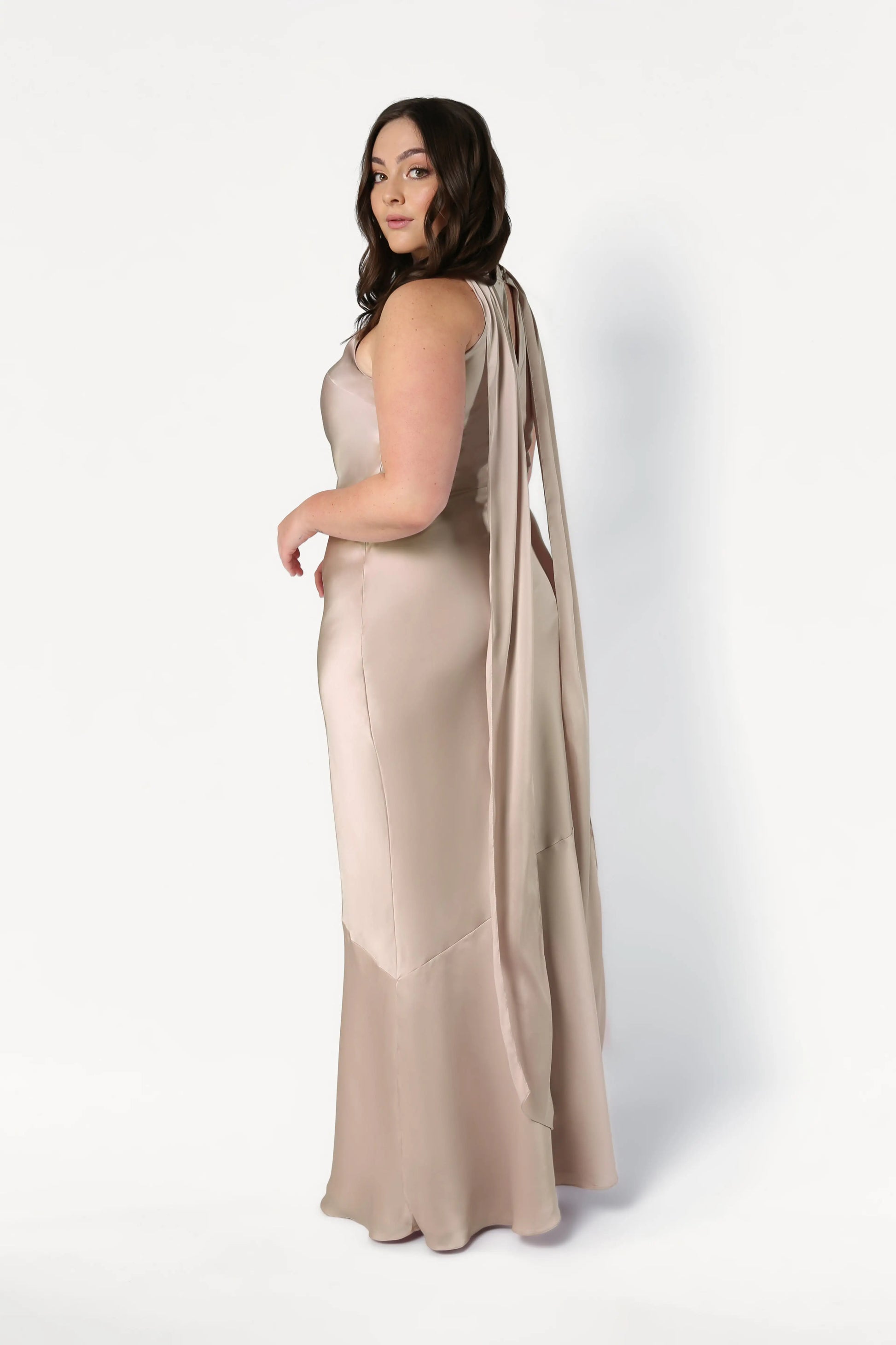 A woman with long brown hair models the TH&TH Mira Bridesmaid Dress in Champagne Sky Satin, a sleeveless, floor-length gown, standing sideways and looking over her shoulder against a plain white background.