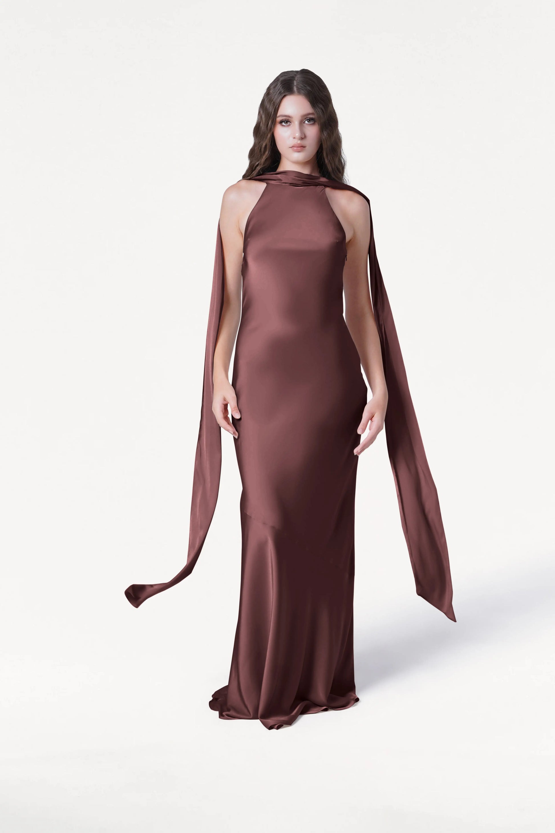 A woman with long brown hair wears the Mira Bridesmaid Dress in Chocolate Brown Satin by TH&TH, featuring sleeveless bias-cut details and long matching scarf panels, standing against a plain white background.