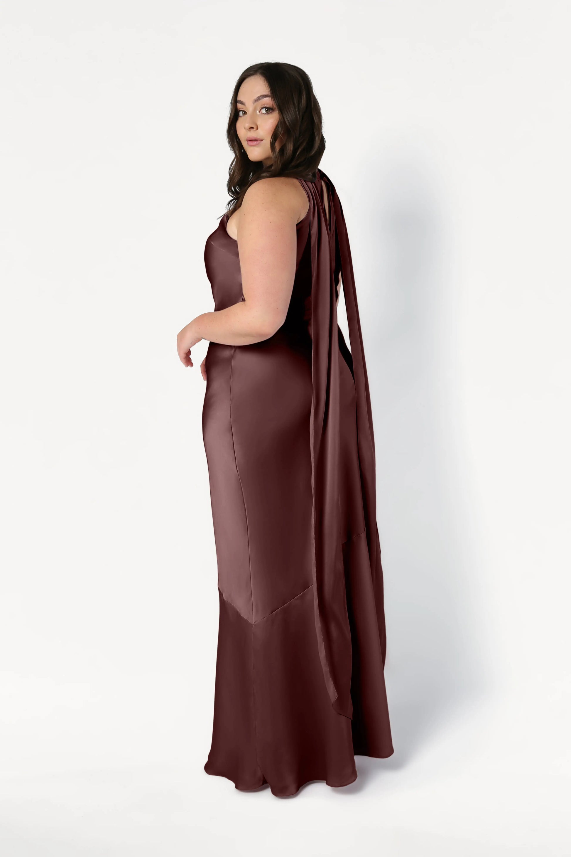 A woman with long, wavy brown hair poses against a white background wearing the TH&TH Mira Bridesmaid Dress in chocolate brown satin, a sleeveless, floor-length gown with a draped sash at the back.