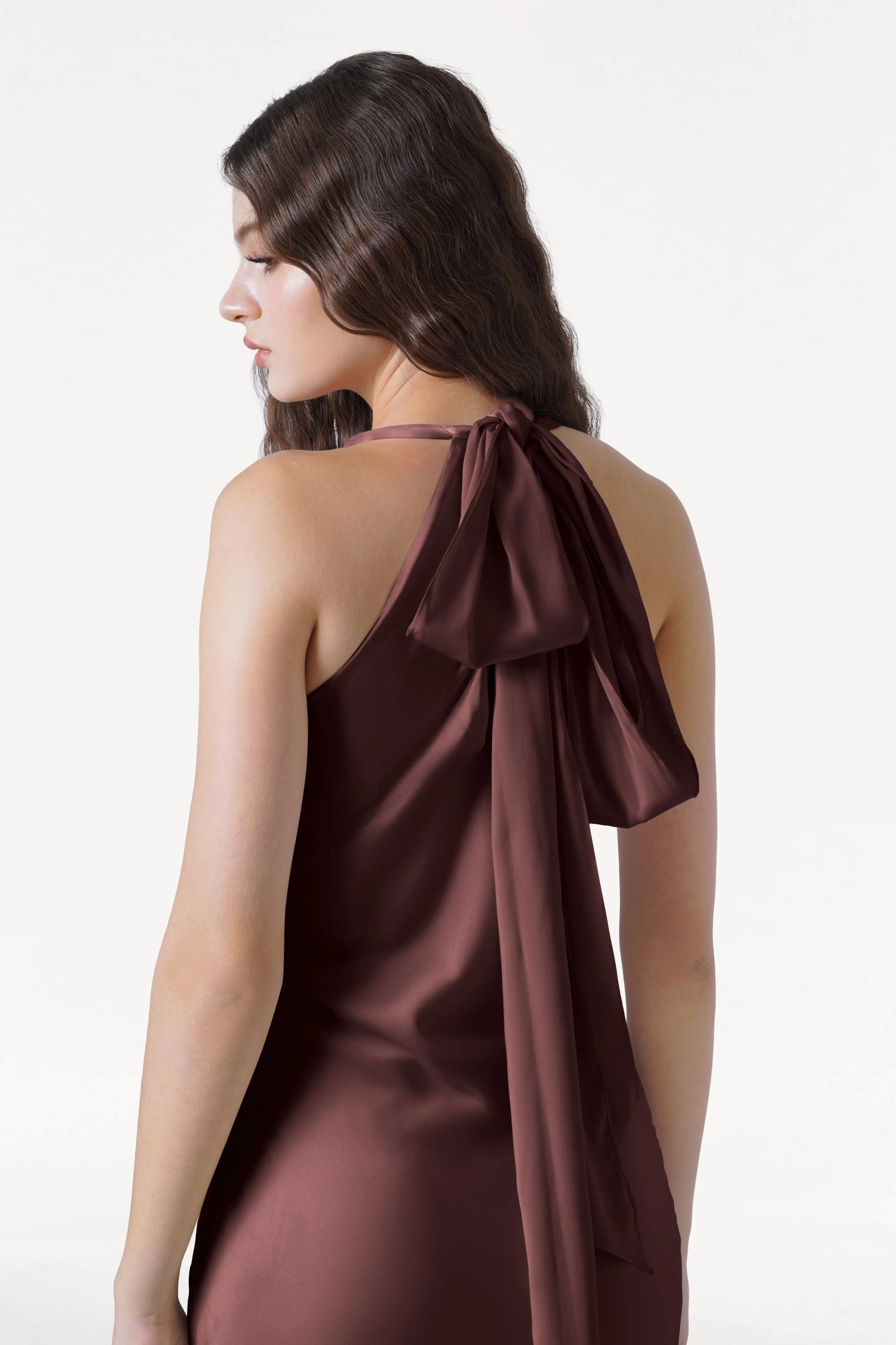 A woman with long, wavy brown hair models the Mira Bridesmaid Dress in Chocolate Brown Satin by TH&TH, featuring a halterneck and large back bow. She stands sideways against a plain white background.