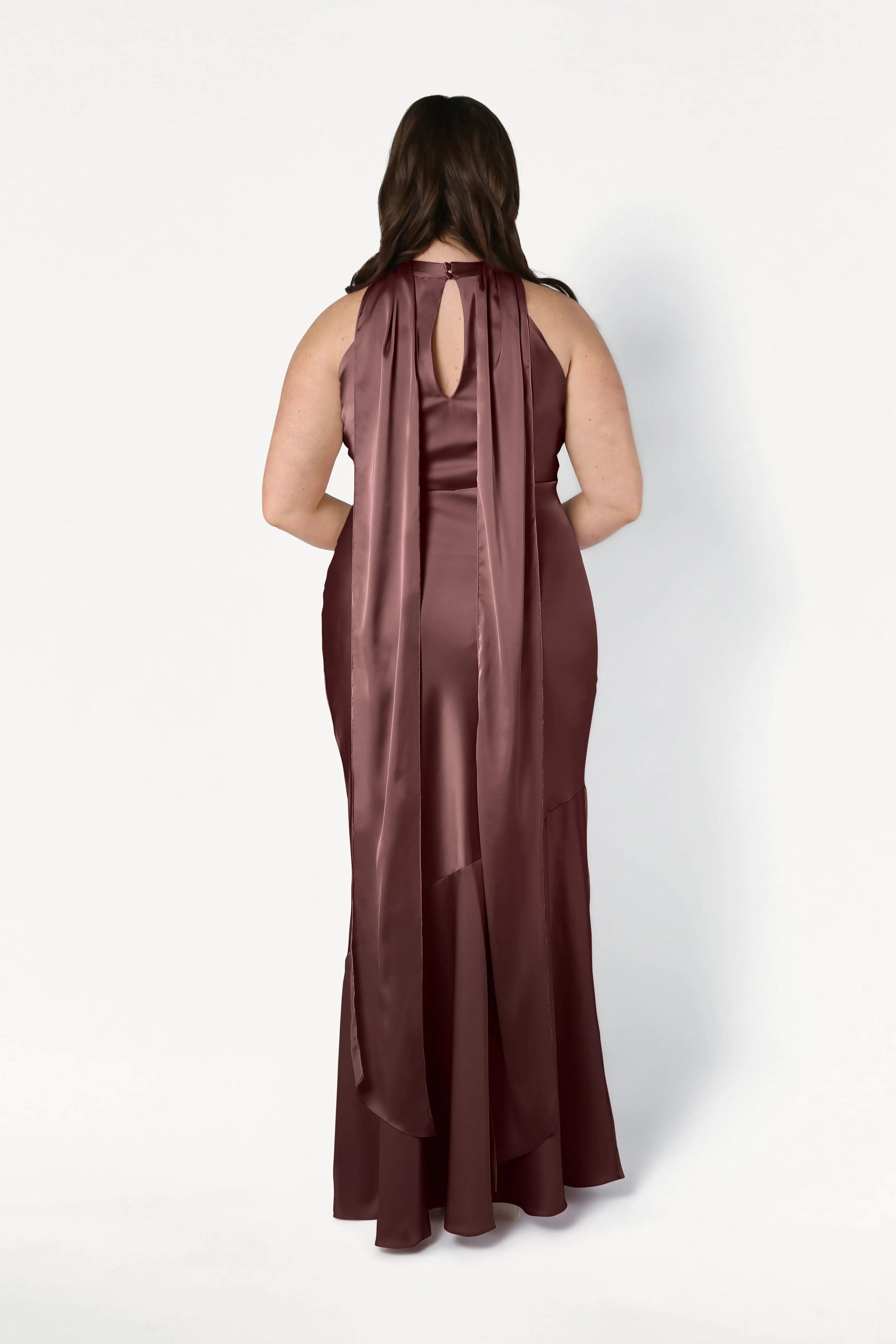 A woman is shown from the back wearing the TH&TH Mira Bridesmaid Dress in chocolate brown satin, a sleeveless, floor-length gown featuring a keyhole neckline and two long, wide trailing ribbons down her back.