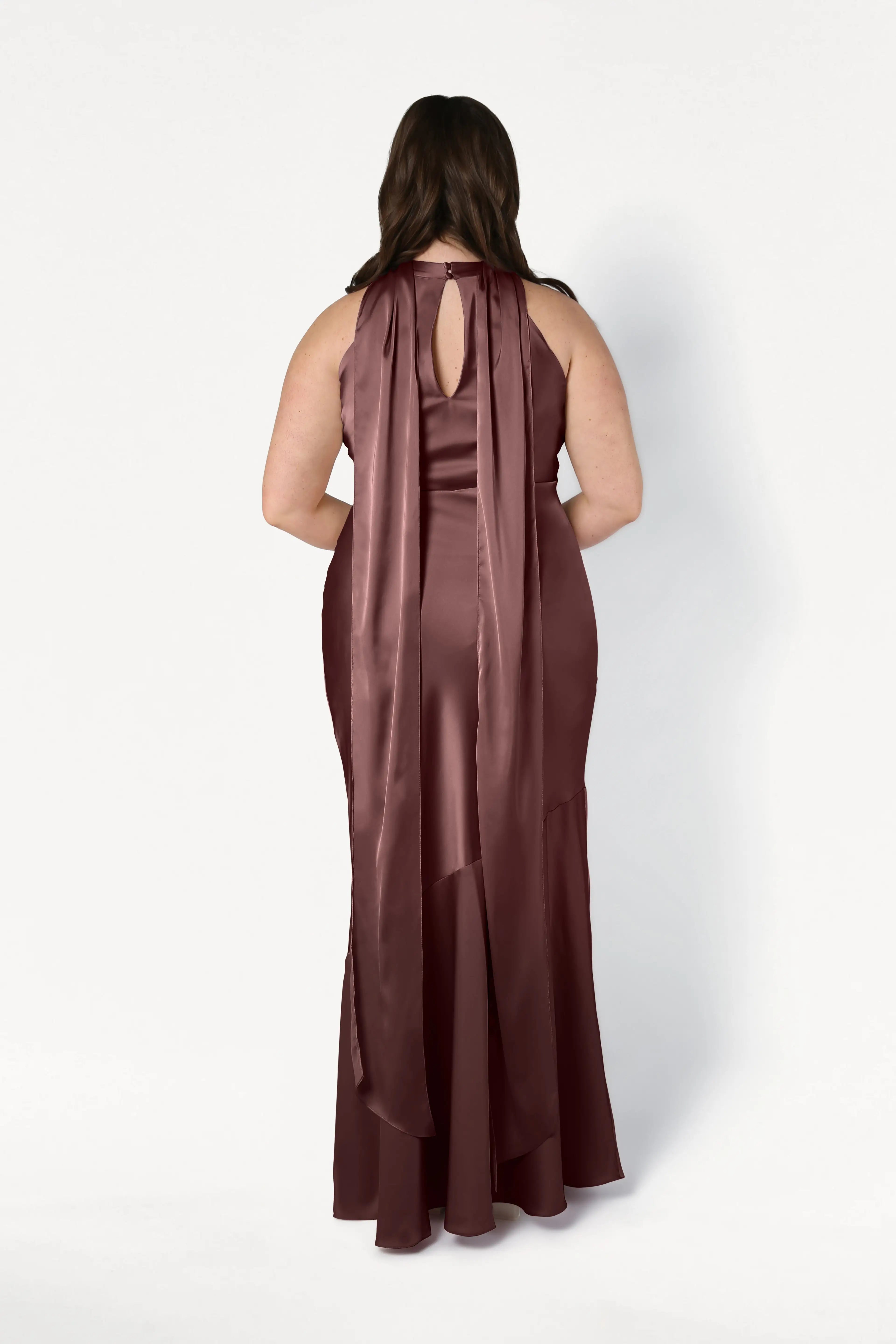 A woman is shown from the back wearing the TH&TH Mira Bridesmaid Dress in chocolate brown satin, a sleeveless, floor-length gown featuring a keyhole neckline and two long, wide trailing ribbons down her back.
