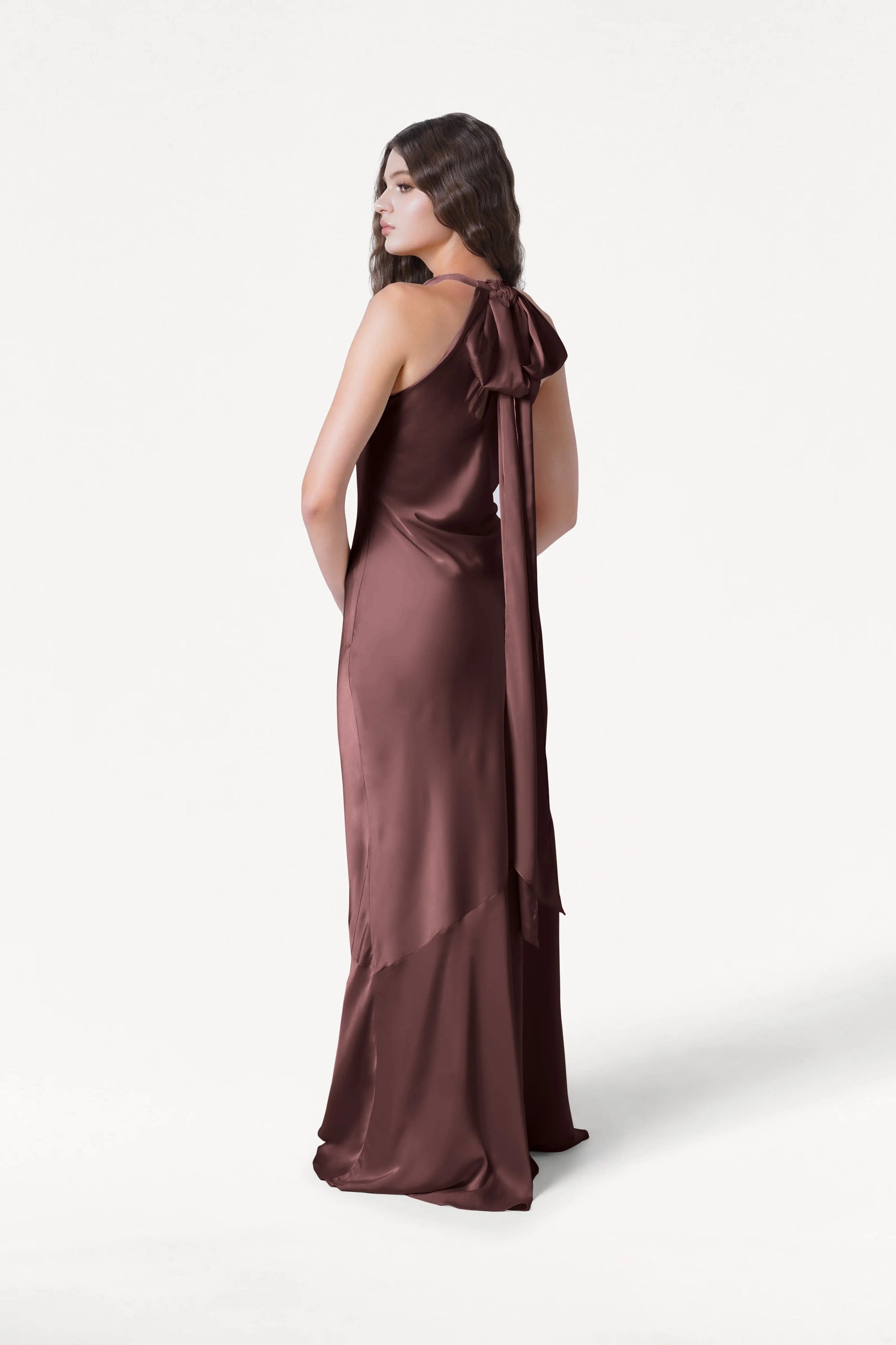 A woman poses with her back turned in the TH&TH Mira Bridesmaid Dress, a floor-length, bias-cut gown in chocolate brown satin with a long ribbon bow at the back. Her long brown hair drapes down against a plain white background.