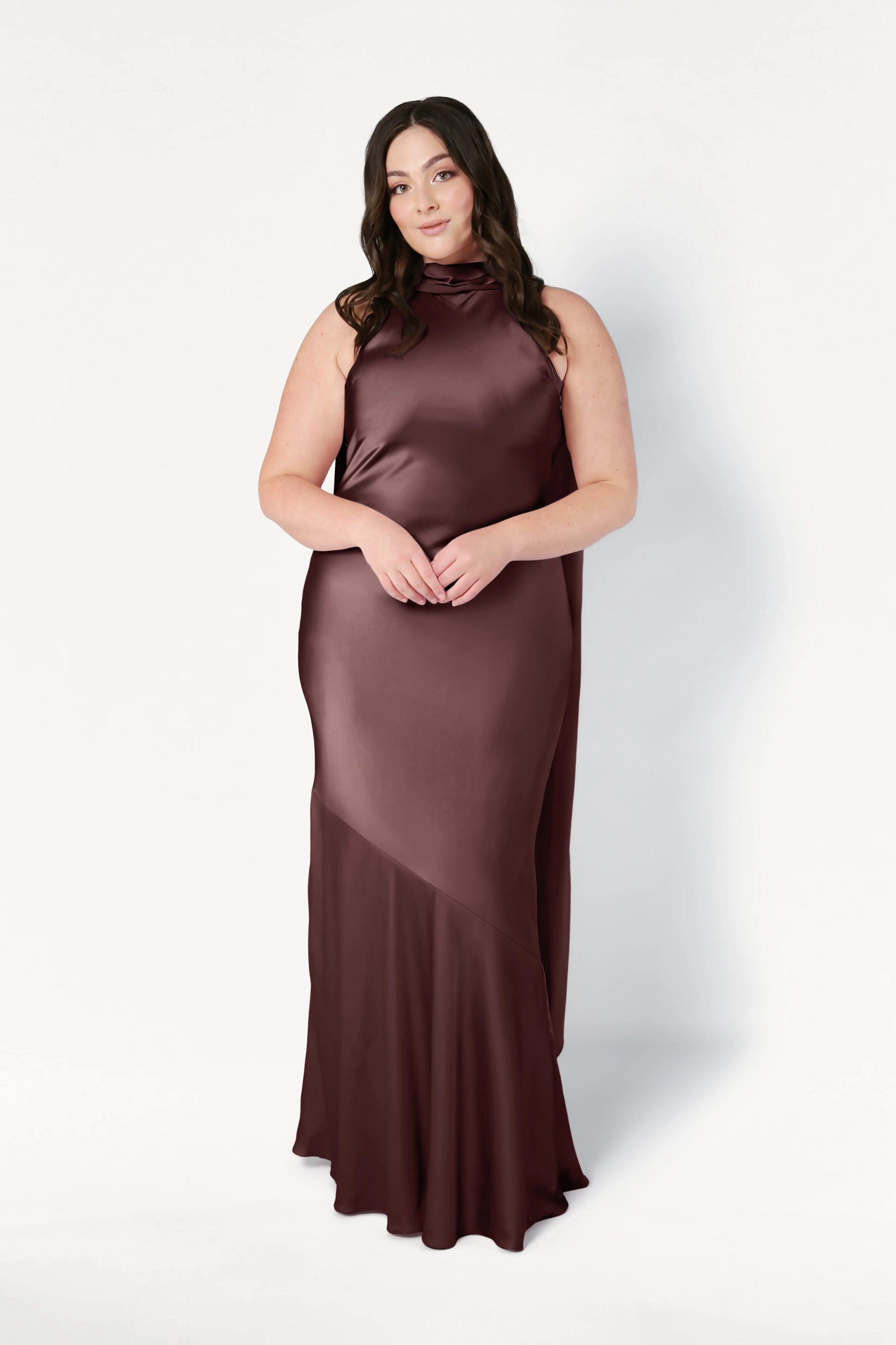 A woman with long, dark hair models the TH&TH Mira Bridesmaid Dress in chocolate brown satin—a sleeveless gown with a high neckline and floor-length hem—against a plain white background.