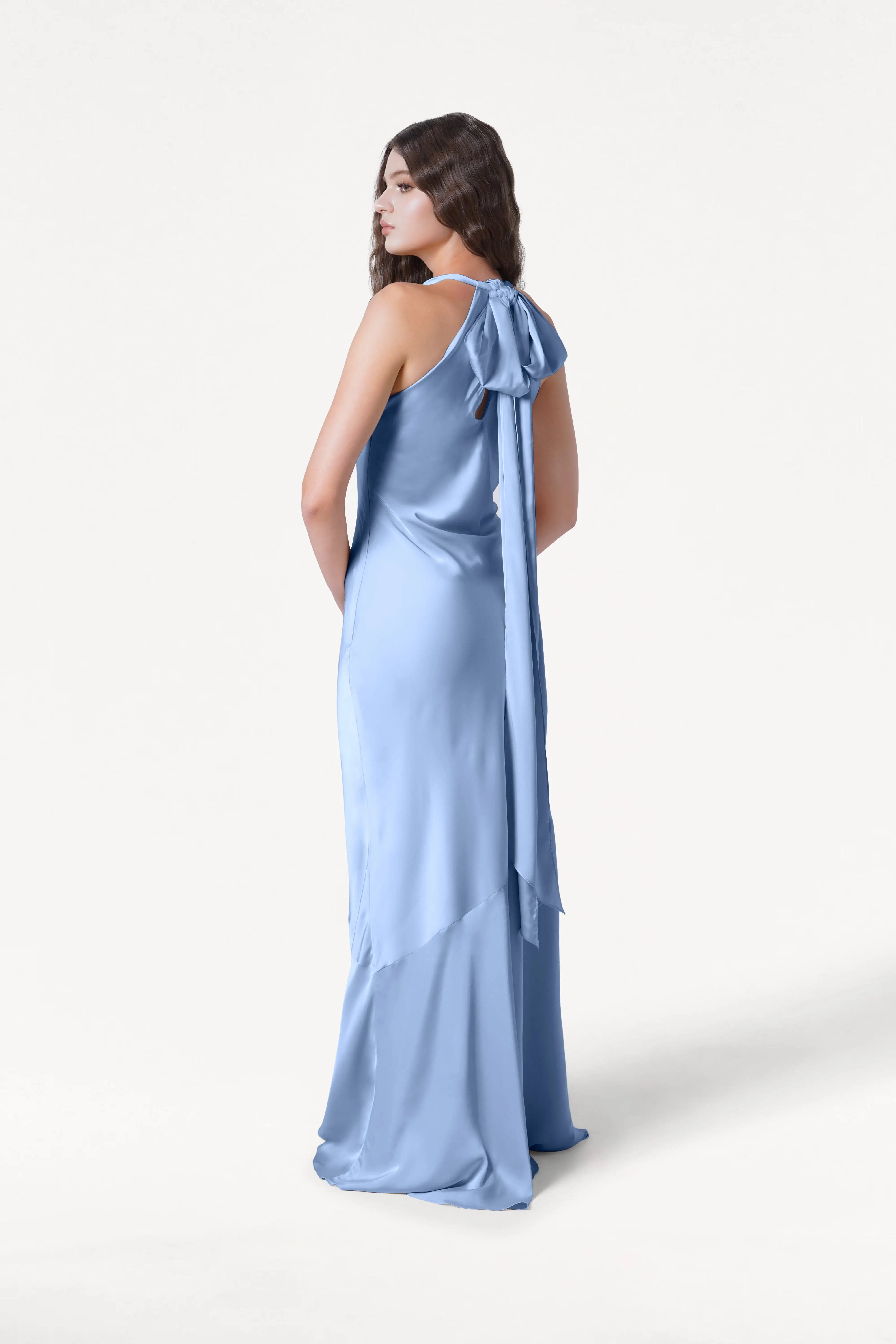 A woman wears the TH&TH Mira Bridesmaid Dress in Dusty Blue Satin, a long, silky bias-cut gown featuring a bow and draped sash at the back, facing away and glancing over her shoulder against a white background.