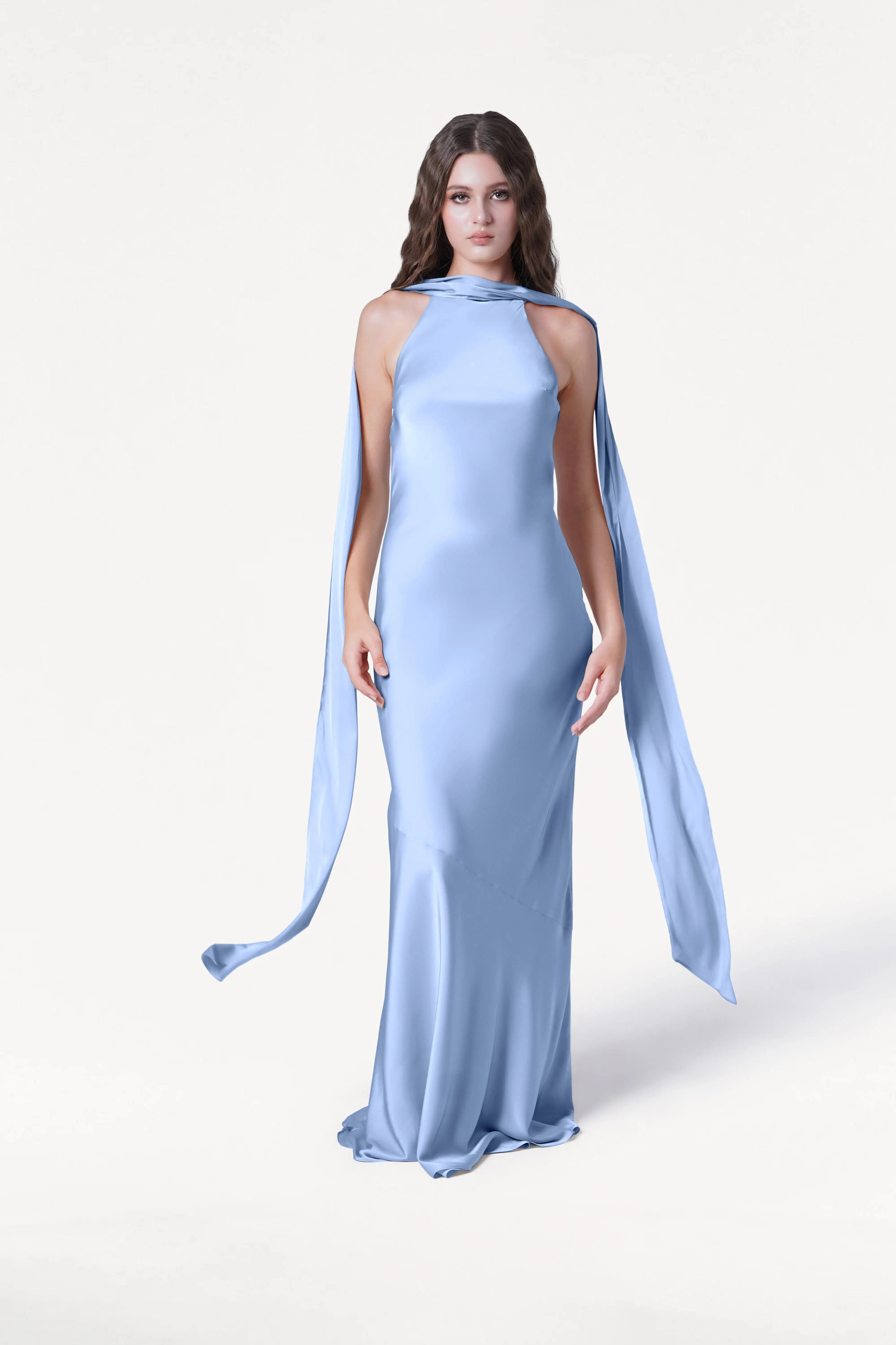 A woman wears the TH&TH Mira Bridesmaid Dress in Dusty Blue Satin, featuring a long silhouette, halterneck, high neckline, and flowing scarf details, standing against a plain white background.