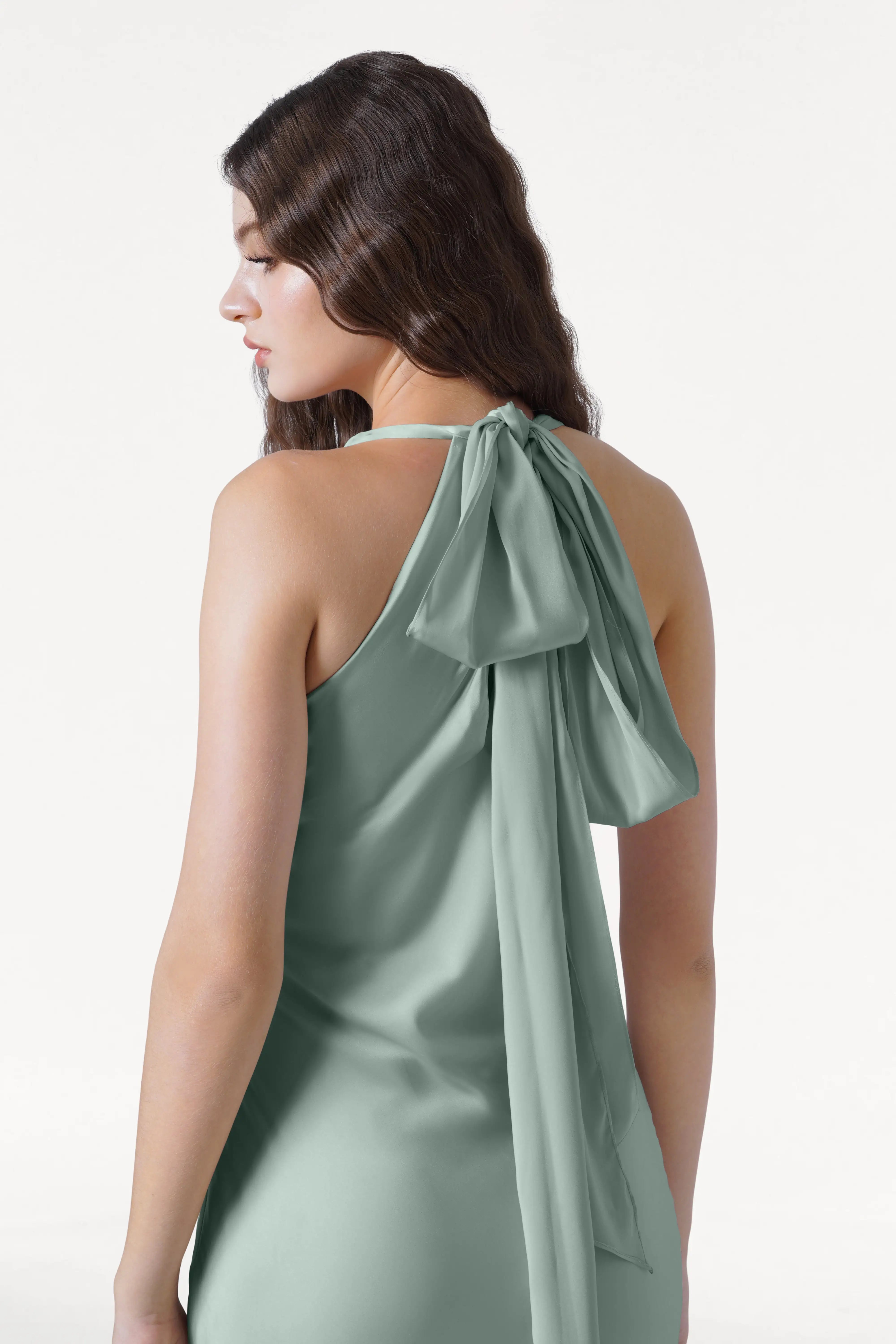 A woman with wavy brown hair is seen from the back, wearing the TH&TH Mira Bridesmaid Dress in Dusty Eucalyptus Satin, a sleeveless halterneck style with a large flowing bow at the neck, set against a plain white background.
