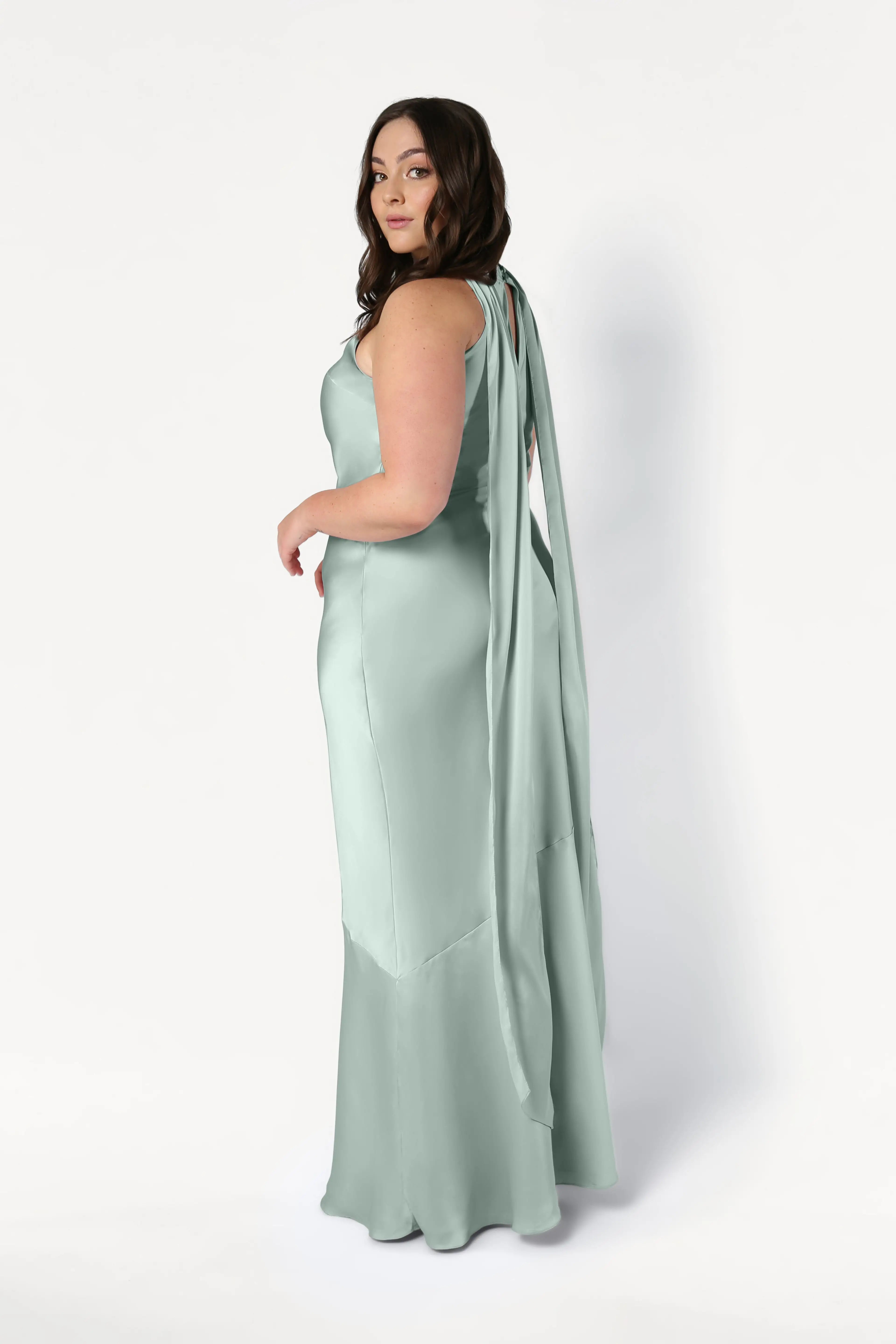 A woman with long brown hair stands sideways against a white background, wearing the TH&TH Mira Bridesmaid Dress in Dusty Eucalyptus Satin, featuring a floor-length hem and long draped straps down the back.