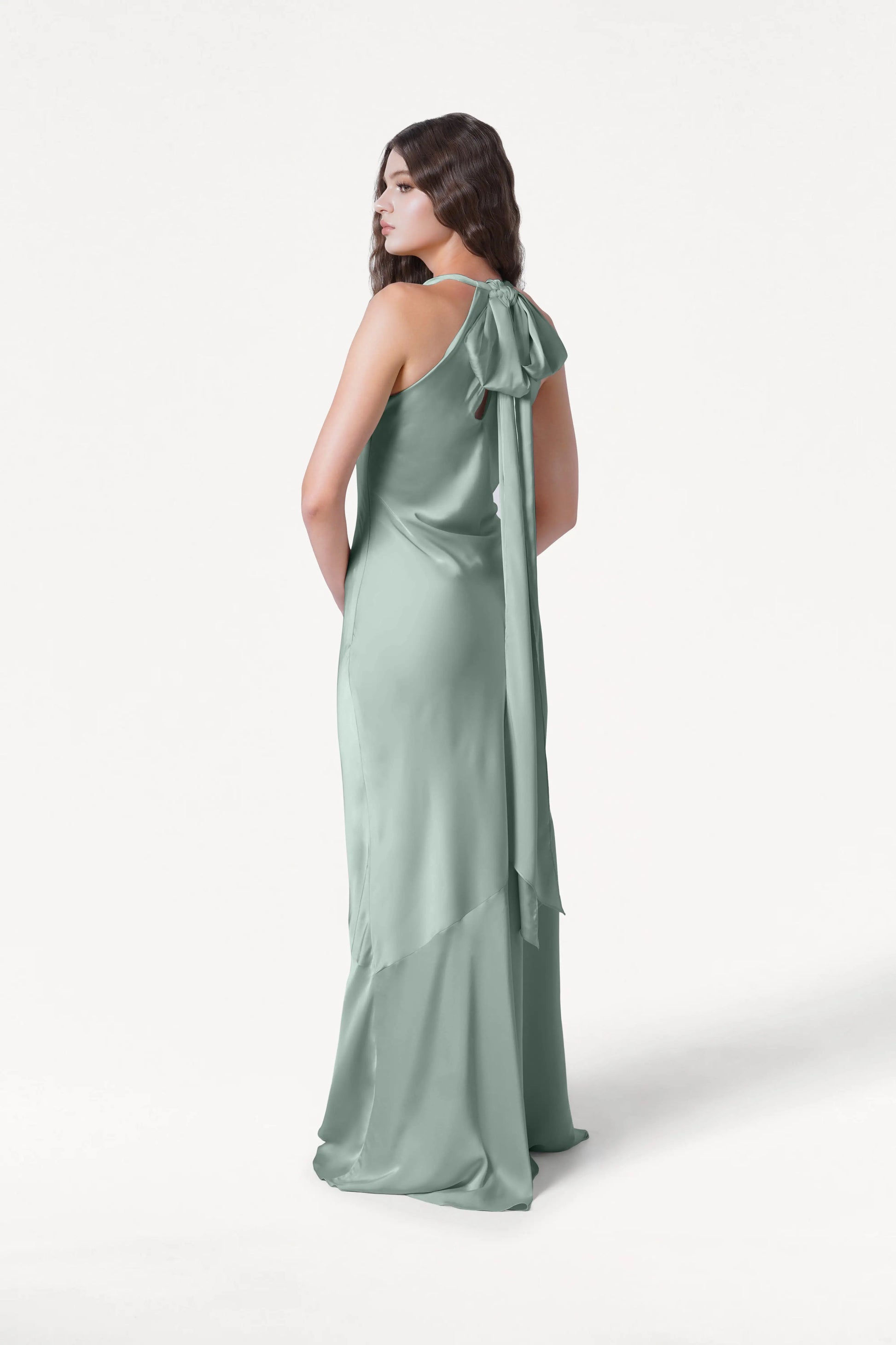 A woman wears the TH&TH Mira Bridesmaid Dress in Dusty Eucalyptus Satin, featuring a silky halterneck and long sash tied at the back, as she stands facing away and looks over her shoulder against a plain white background.