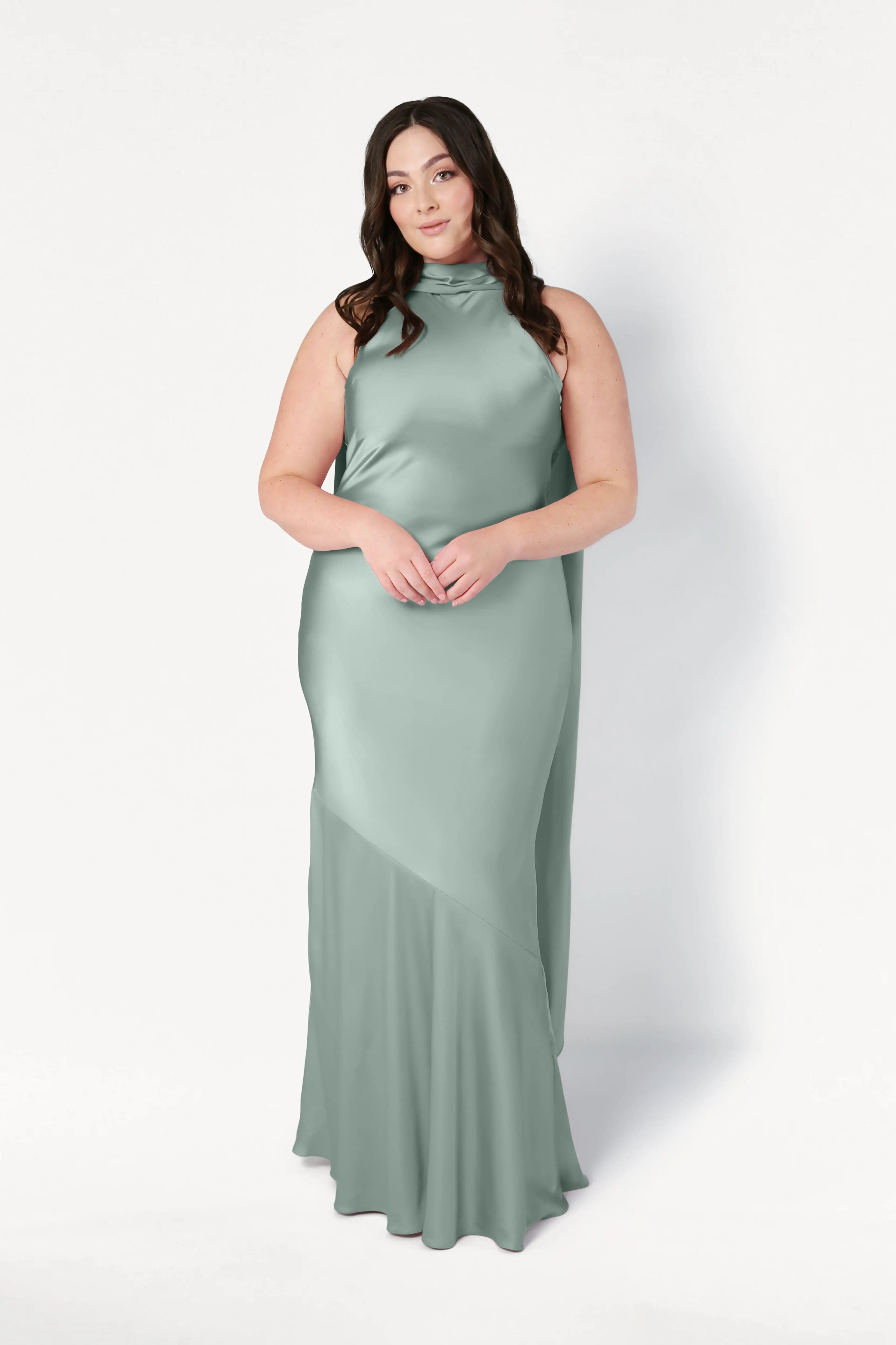 A woman with long, dark hair wears the TH&TH Mira Bridesmaid Dress in Dusty Eucalyptus Satin, featuring a sleeveless design and high neckline, standing against a plain white background.