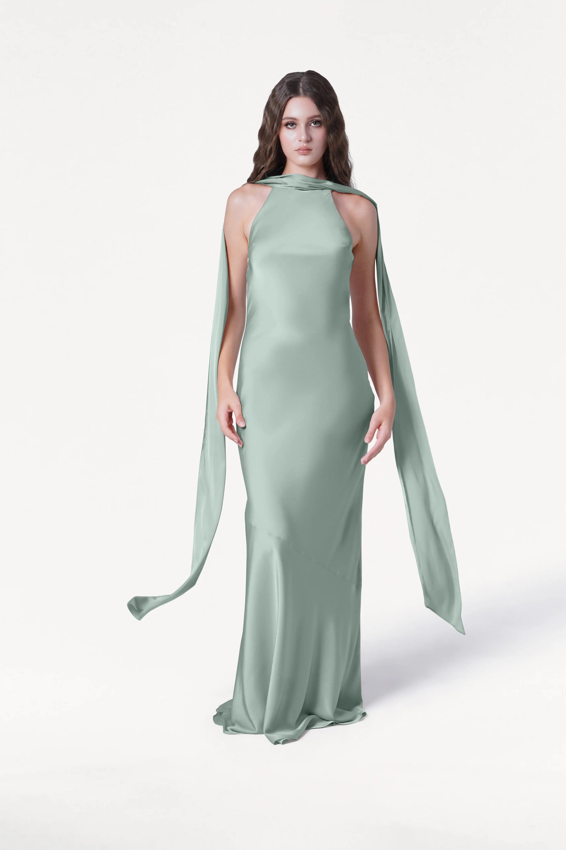 A woman with long brown hair wears the TH&TH Mira Bridesmaid Dress in Dusty Eucalyptus Satin, featuring a sleek halterneck and flowing shoulder accents, standing against a plain white background.
