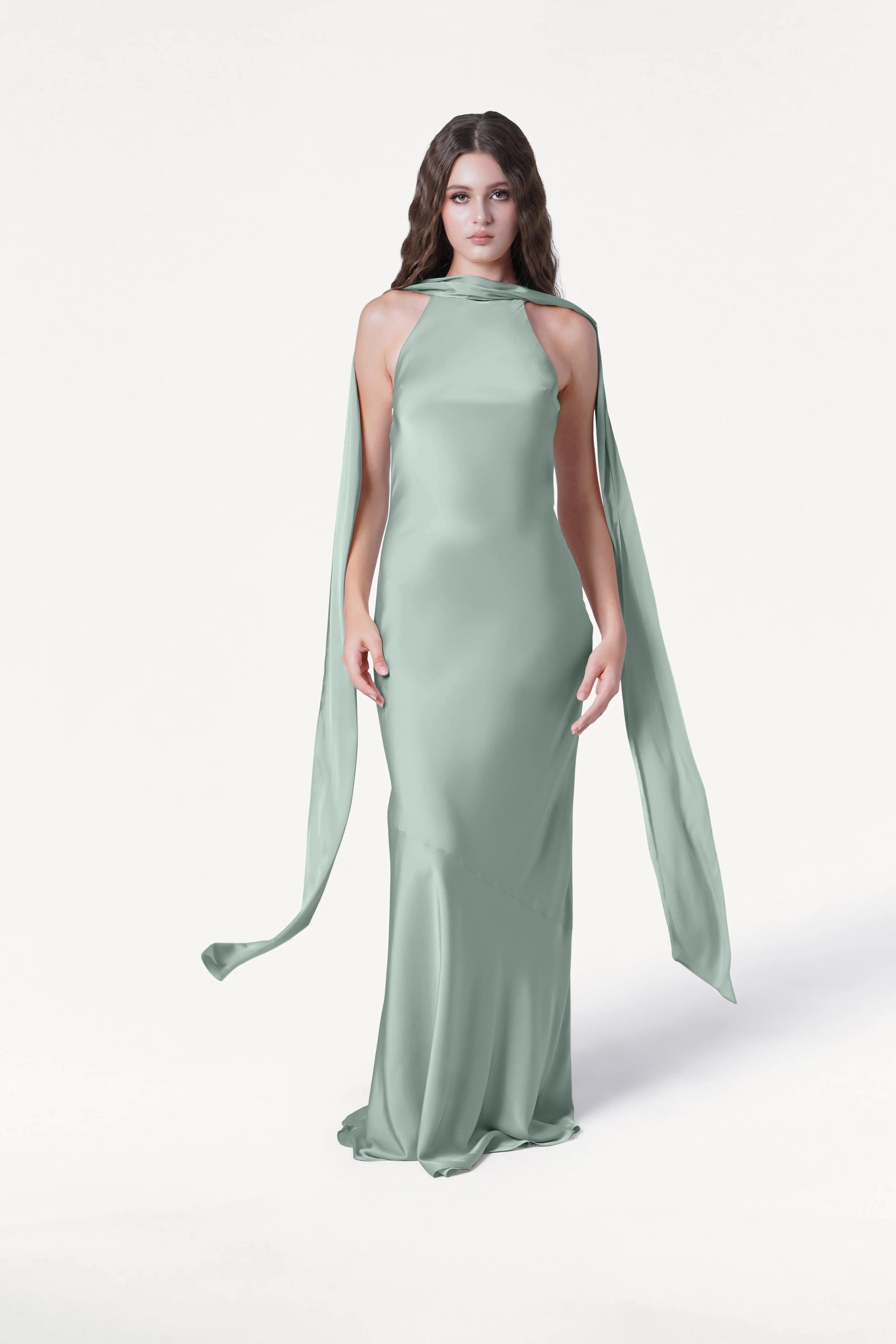 A woman with long brown hair wears the TH&TH Mira Bridesmaid Dress in Dusty Eucalyptus Satin, featuring a sleek halterneck and flowing shoulder accents, standing against a plain white background.