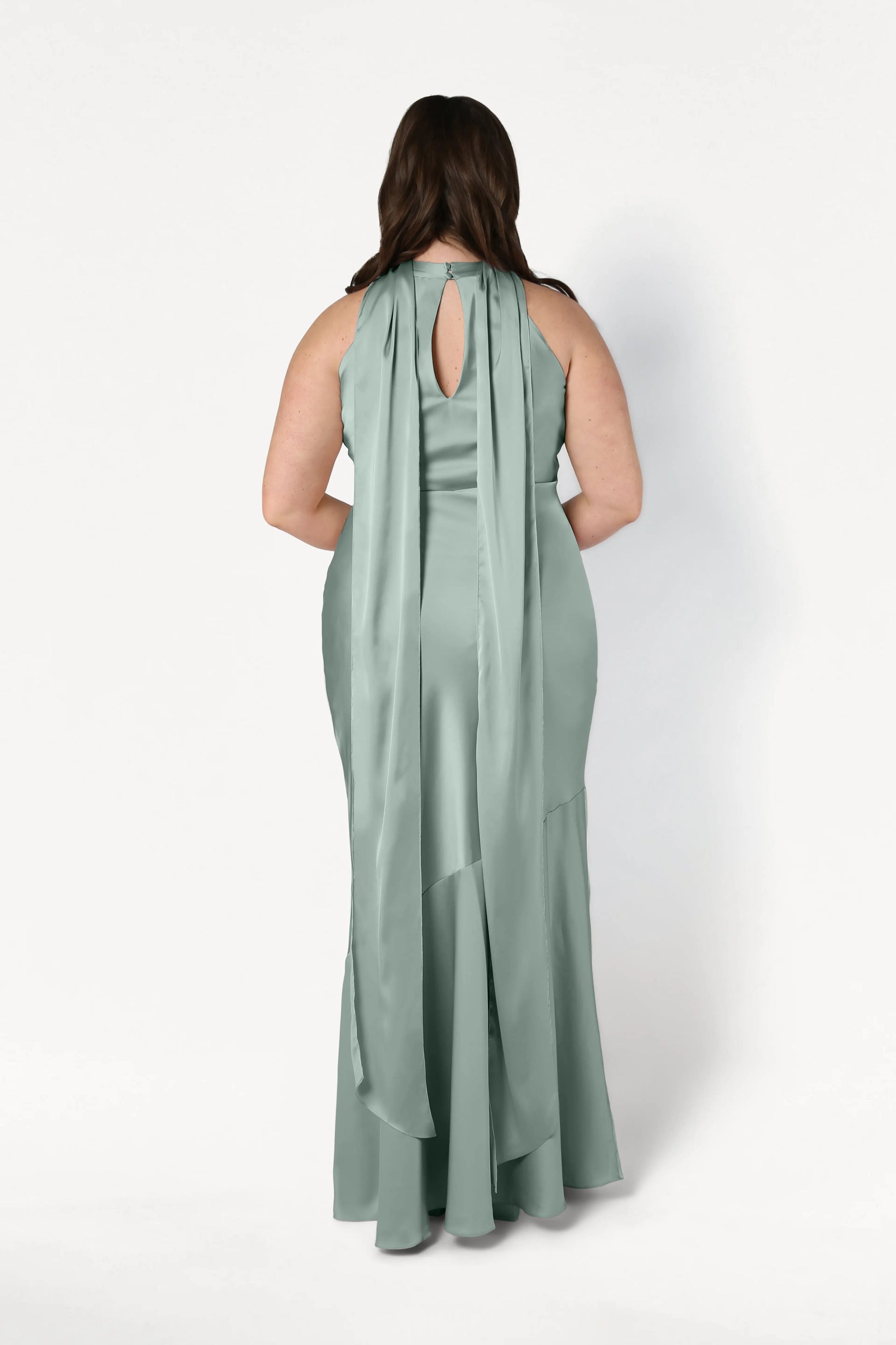 A woman wears the TH&TH Mira Bridesmaid Dress in Dusty Eucalyptus Satin, featuring a bias-cut silhouette, long draped scarf-like details from the shoulders, and a keyhole opening at the upper back.