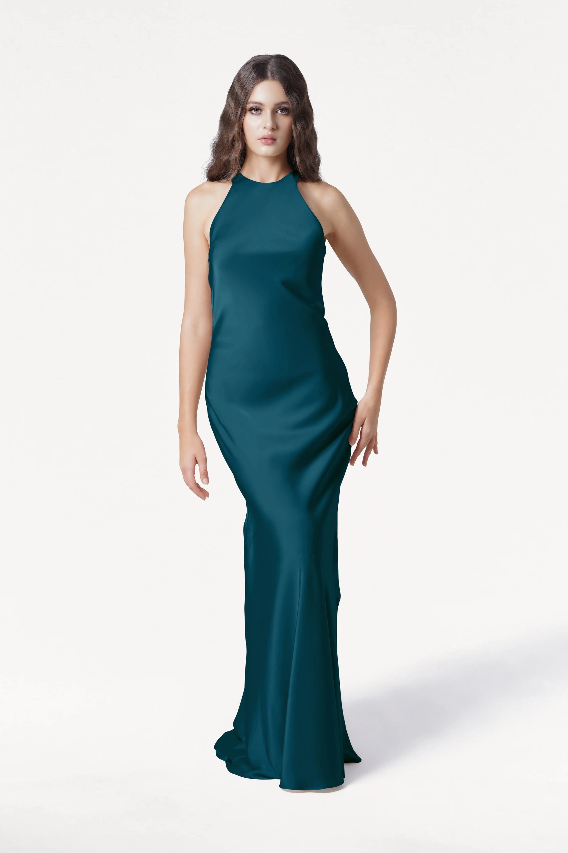 A woman with long wavy hair models the TH&TH Mira Bridesmaid Dress in shimmering Emerald Dawn satin, a sleeveless floor-length gown, set against a plain white backdrop.