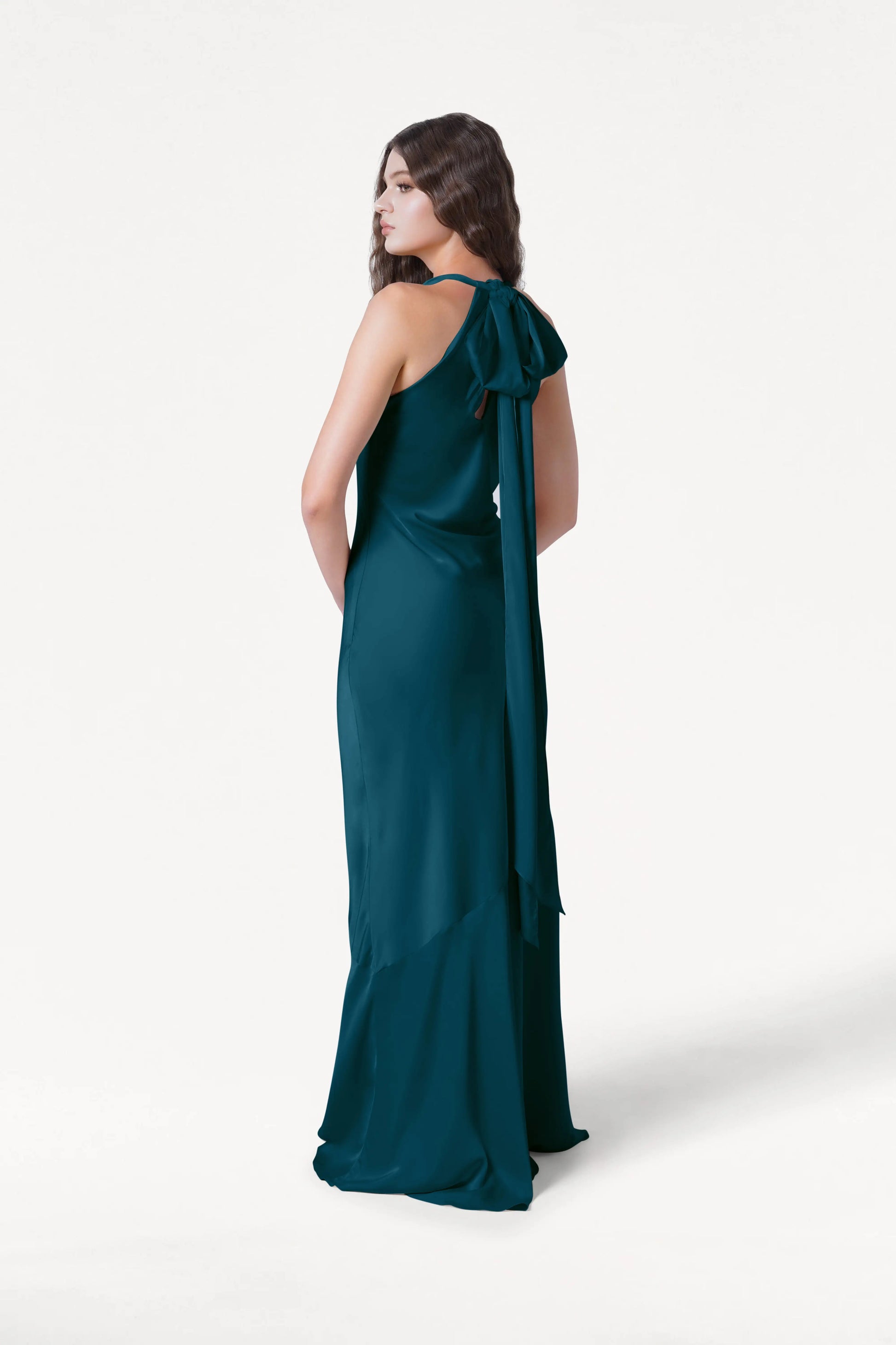 A woman stands in profile wearing the TH&TH Mira Bridesmaid Dress in Emerald Dawn Satin, featuring a long silhouette with a large bow and trailing ribbons at the back, set against a plain white background.