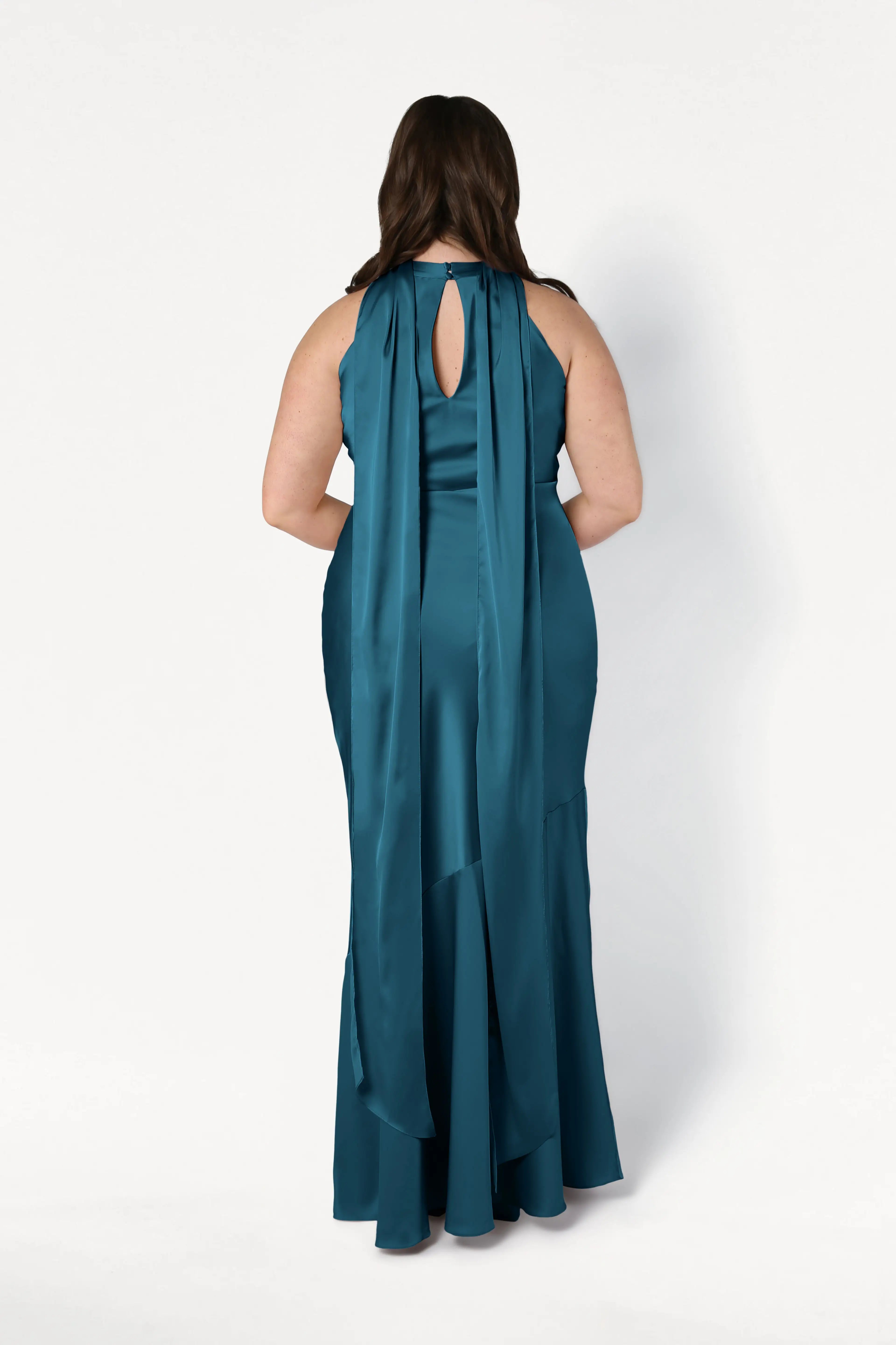 A woman with long brown hair faces away in the TH&TH Mira Bridesmaid Dress in Emerald Dawn Satin, a floor-length gown with a keyhole back and two scarf-like sashes, set against a plain white background.