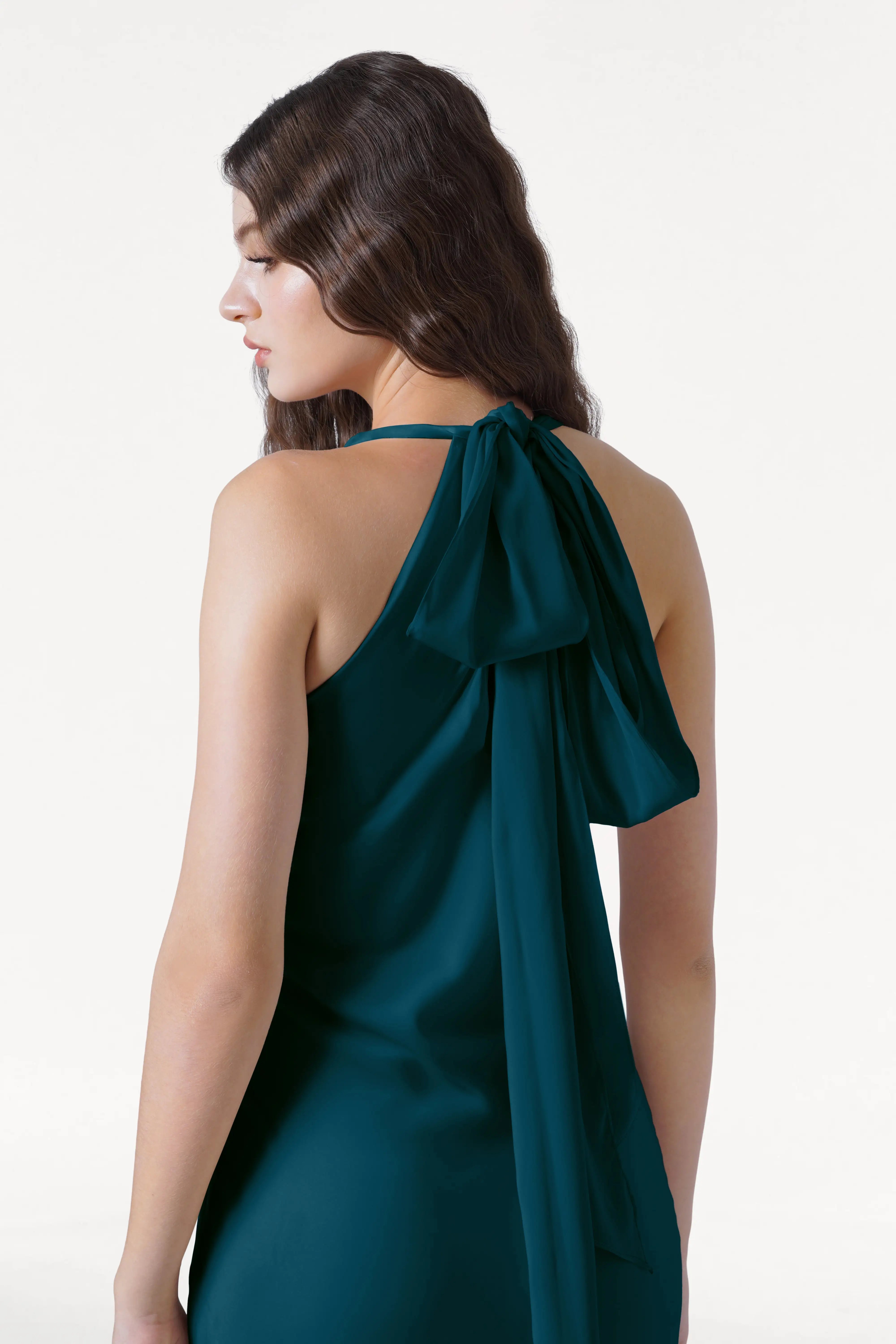 A woman with long wavy brown hair is shown from the side wearing the TH&TH Mira Bridesmaid Dress in Emerald Dawn Satin, featuring a halterneck, large back bow, and draped detail, set against a plain white background.