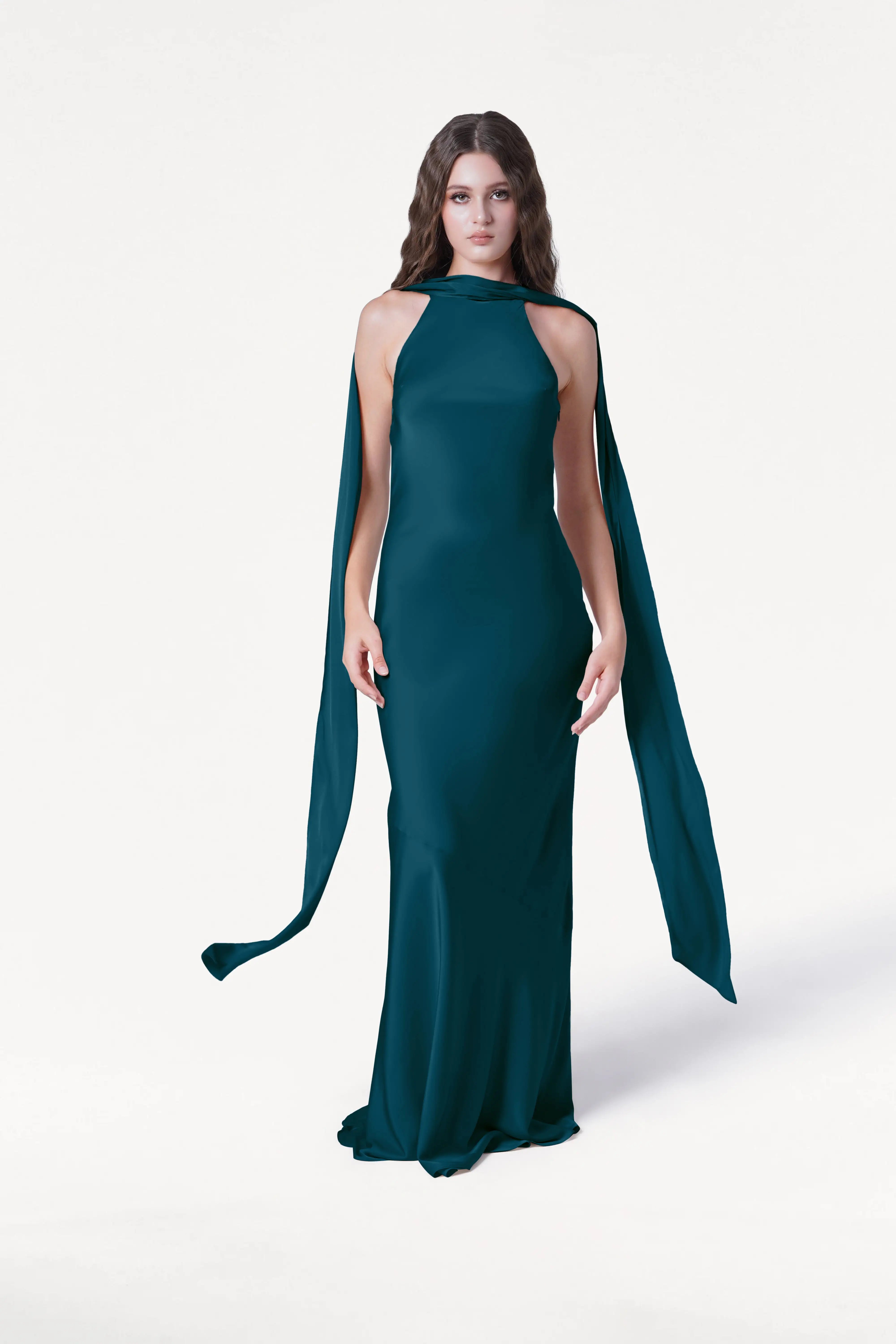 A woman with long wavy brown hair wears the TH&TH Mira Bridesmaid Dress in Emerald Dawn Satin, a sleeveless, floor-length gown with flowing shoulder cape detailing, standing against a plain white background.
