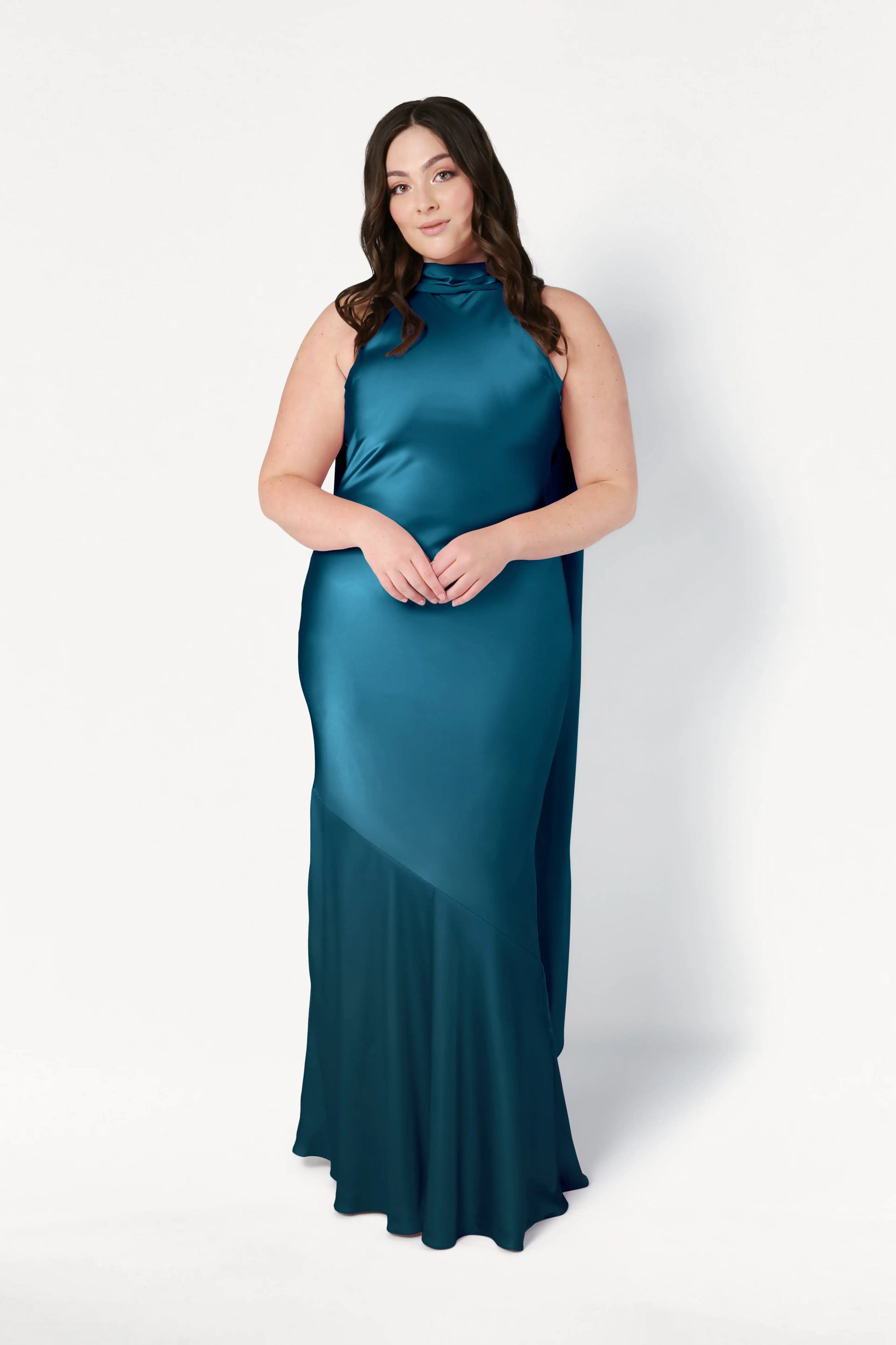 A woman with long brown hair stands against a white background, wearing the TH&TH Mira Bridesmaid Dress in Emerald Dawn Satin—a sleeveless, floor-length gown with a high neckline and asymmetrical hem—her hands gently clasped in front.