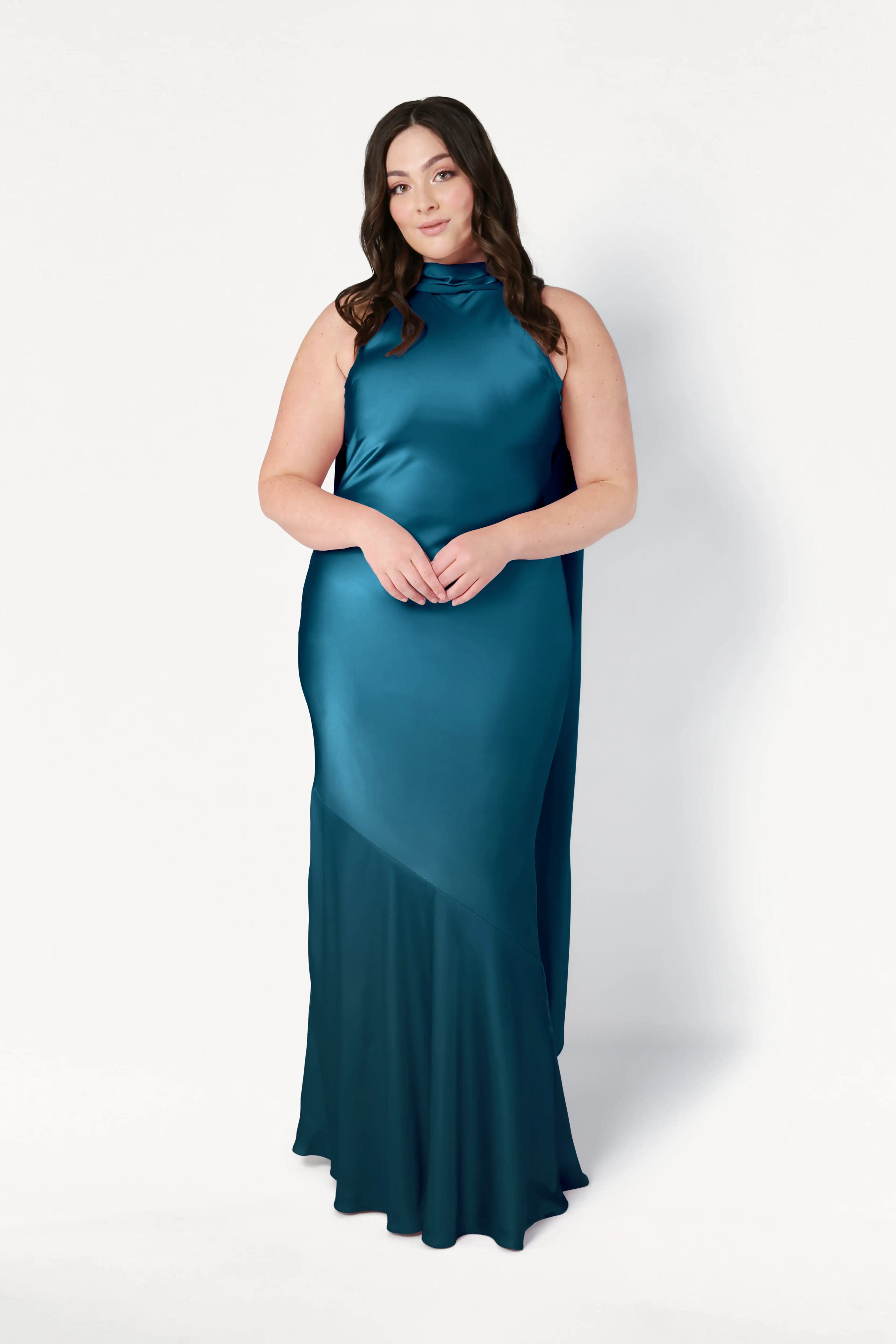 A woman with long brown hair stands against a white background, wearing the TH&TH Mira Bridesmaid Dress in Emerald Dawn Satin—a sleeveless, floor-length gown with a high neckline and asymmetrical hem—her hands gently clasped in front.