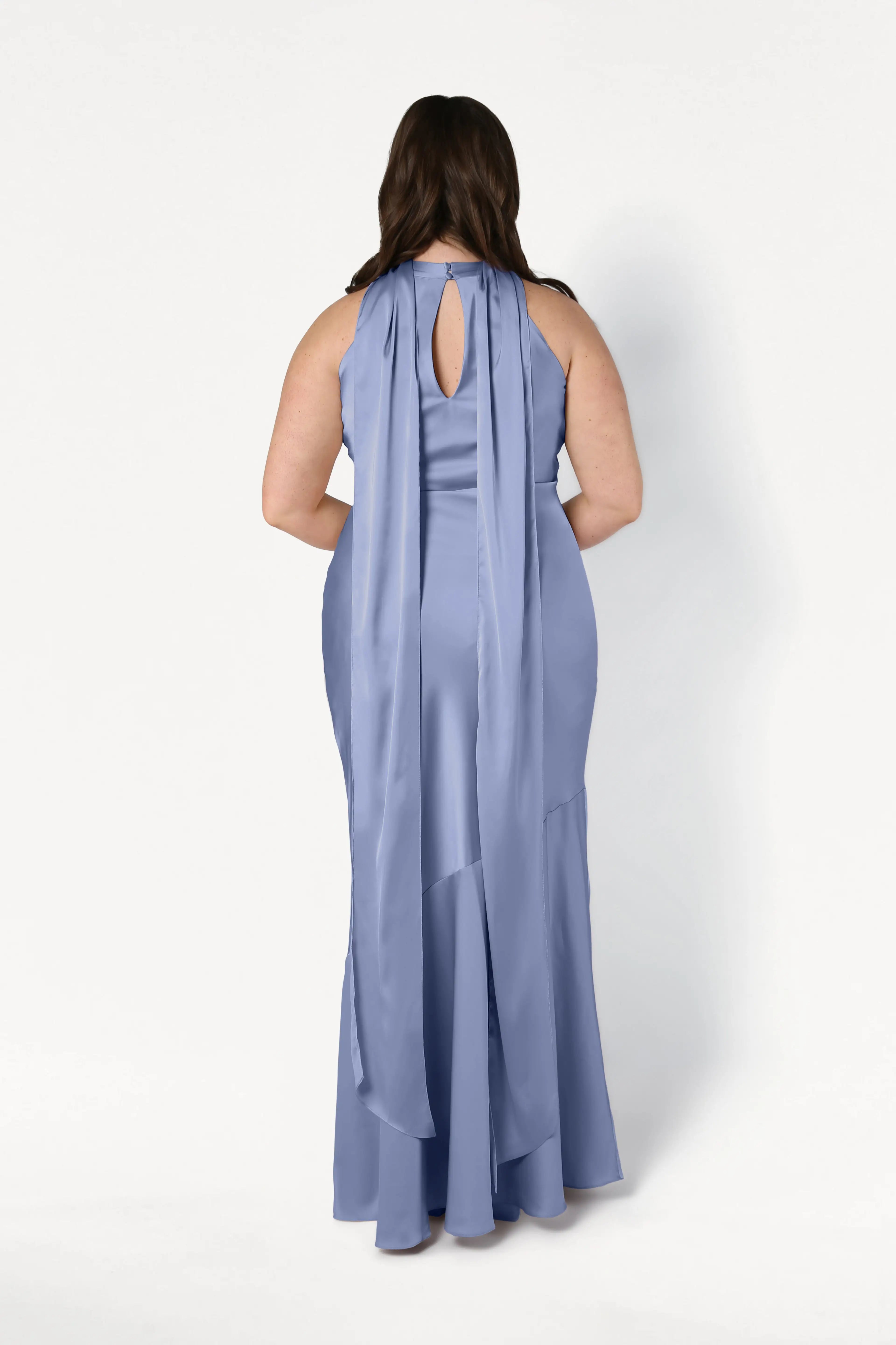 A woman with long brown hair stands with her back to the camera, wearing the TH&TH Mira Bridesmaid Dress in French Blue Satin—sleeveless, floor-length, with a keyhole cutout and two long sashes draping elegantly down the back.