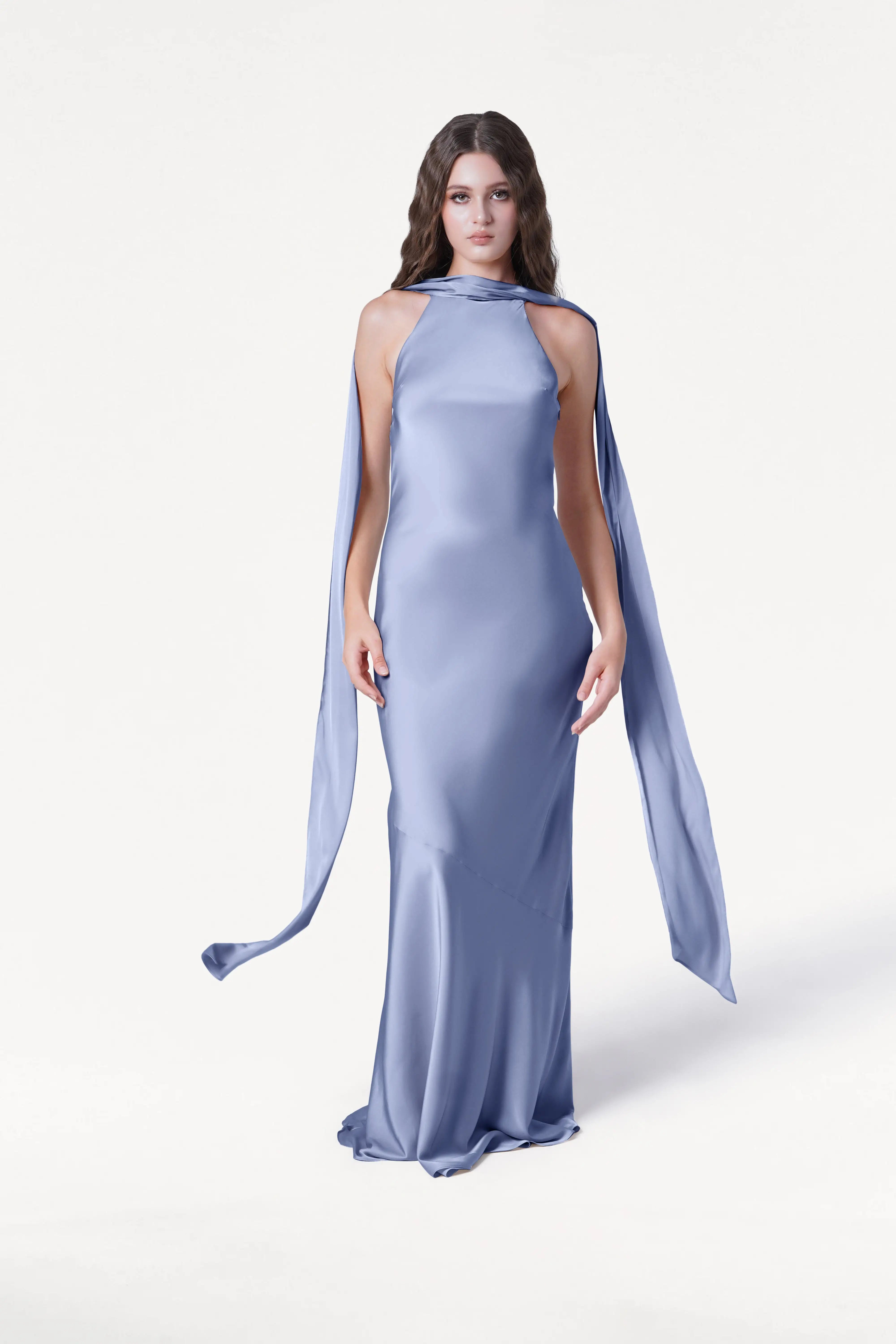 A woman models the TH&TH Mira Bridesmaid Dress in French Blue Satin, featuring a high neckline and long, scarf-like extensions; the elegant, floor-length gown stands out against a plain white background.