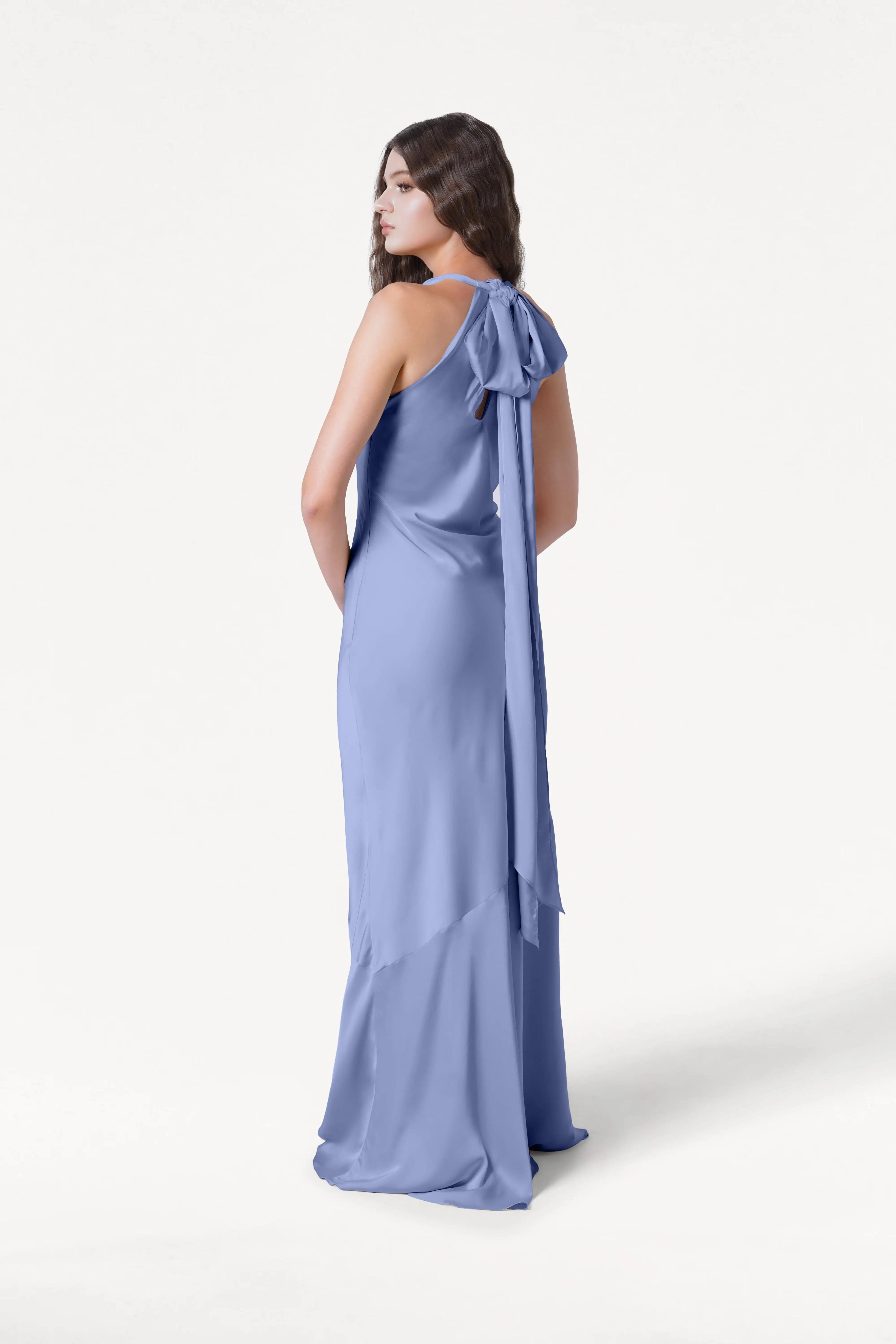 A woman stands sideways in the TH&TH Mira Bridesmaid Dress, a long, French Blue satin gown with a halterneck and a large bow with flowing sash tied at the back. The background is plain white.