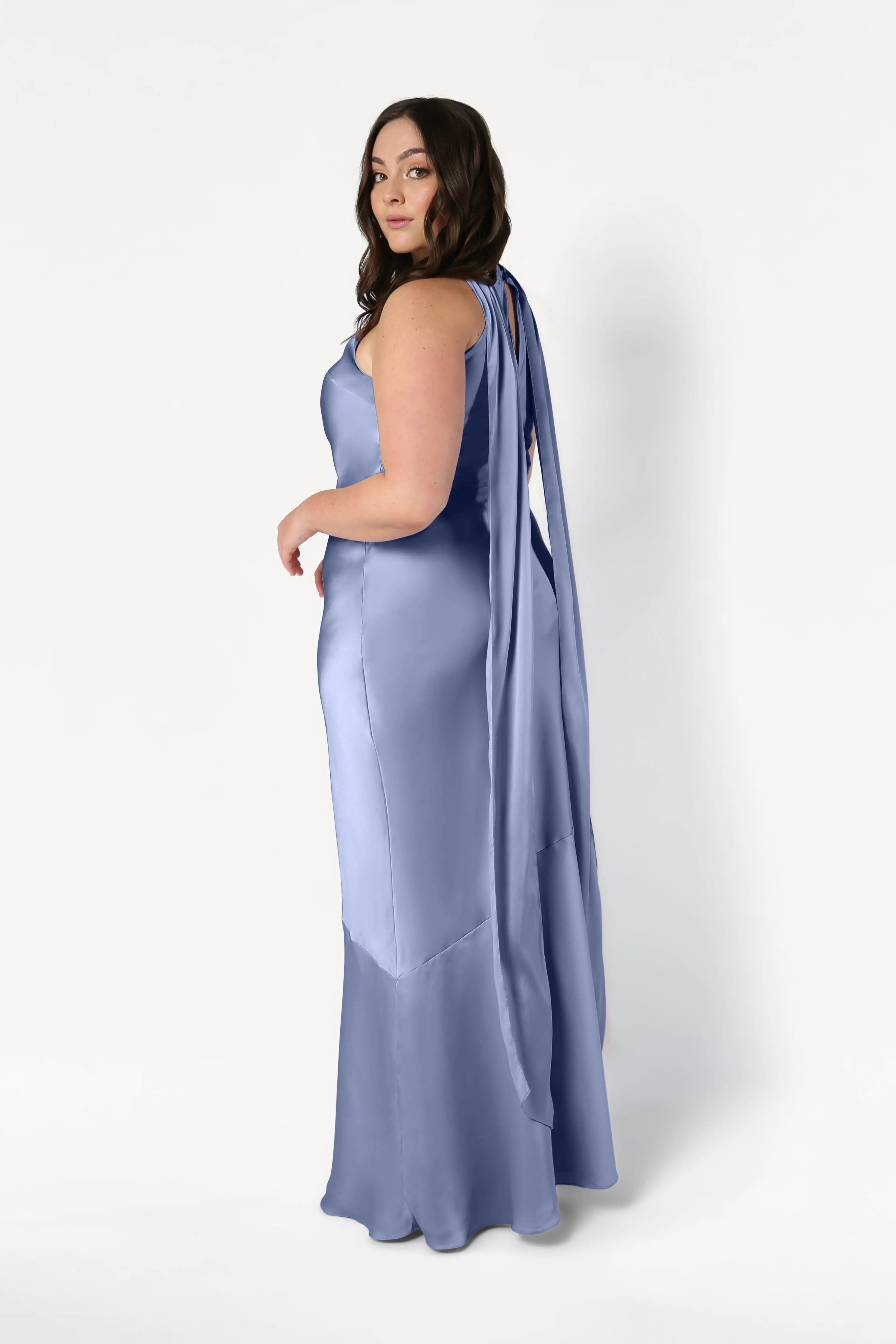 A woman with long brown hair wears the TH&TH Mira Bridesmaid Dress in French Blue Satin, featuring a sleeveless, floor-length silhouette and flowing ribbon back details, as she poses against a plain white background.