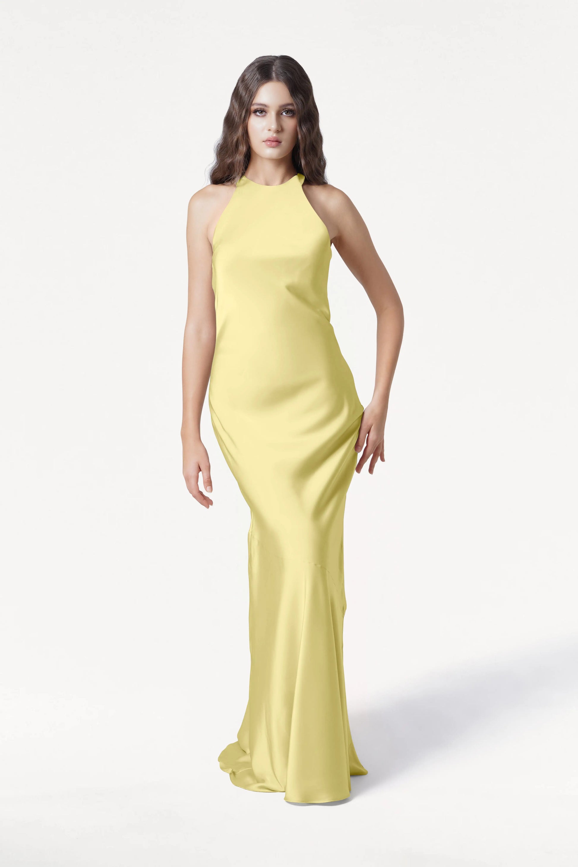 A woman with long brown hair in loose waves and a neutral expression wears the TH&TH Mira Bridesmaid Dress in Limoncello Satin, a sleeveless, floor-length halterneck gown, standing against a white background.