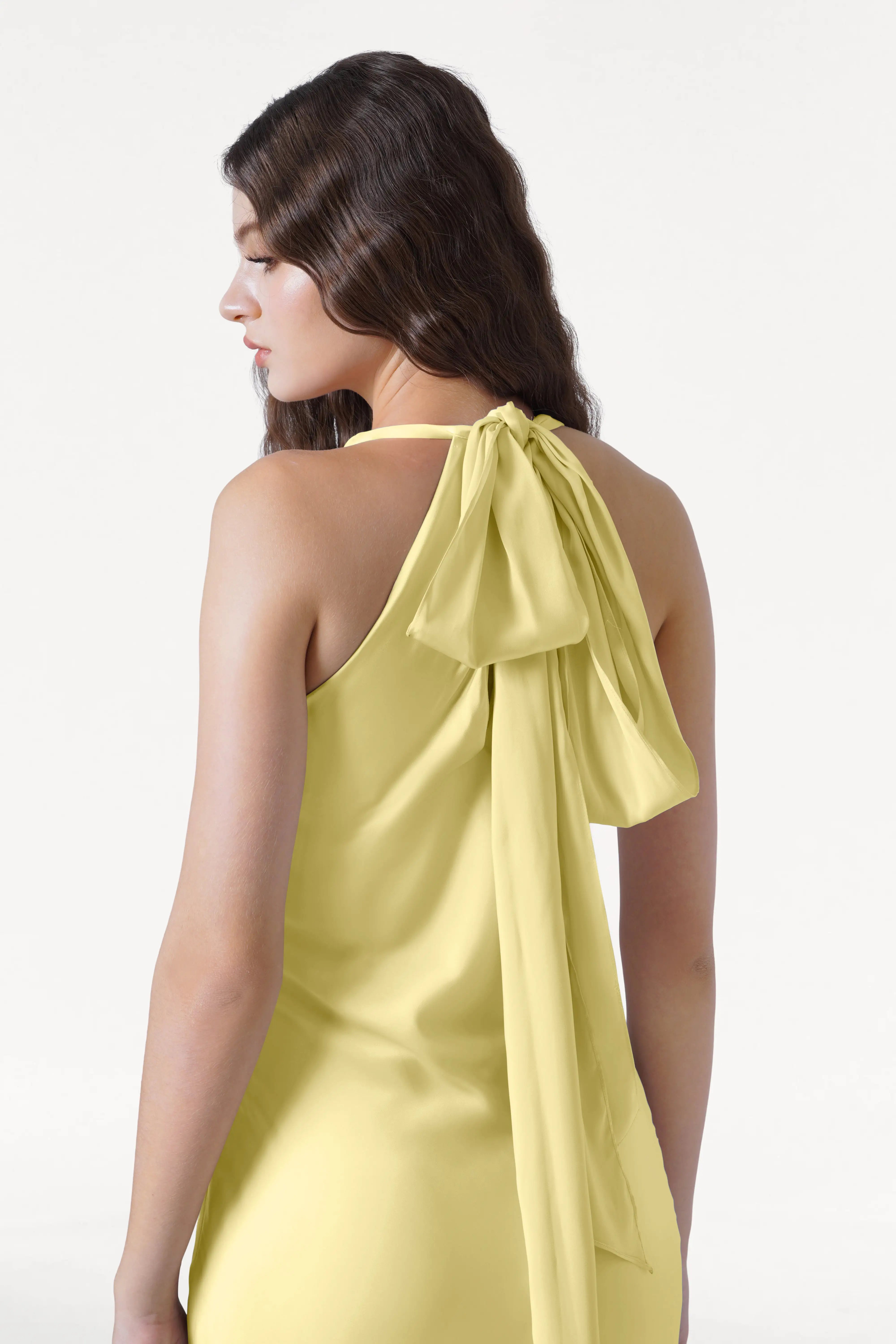 A woman with long wavy brown hair is seen from behind, wearing the TH&TH Mira Bridesmaid Dress in Limoncello Satin, a sleeveless design featuring a large bow and long sash at the back. She looks slightly left against a white background.