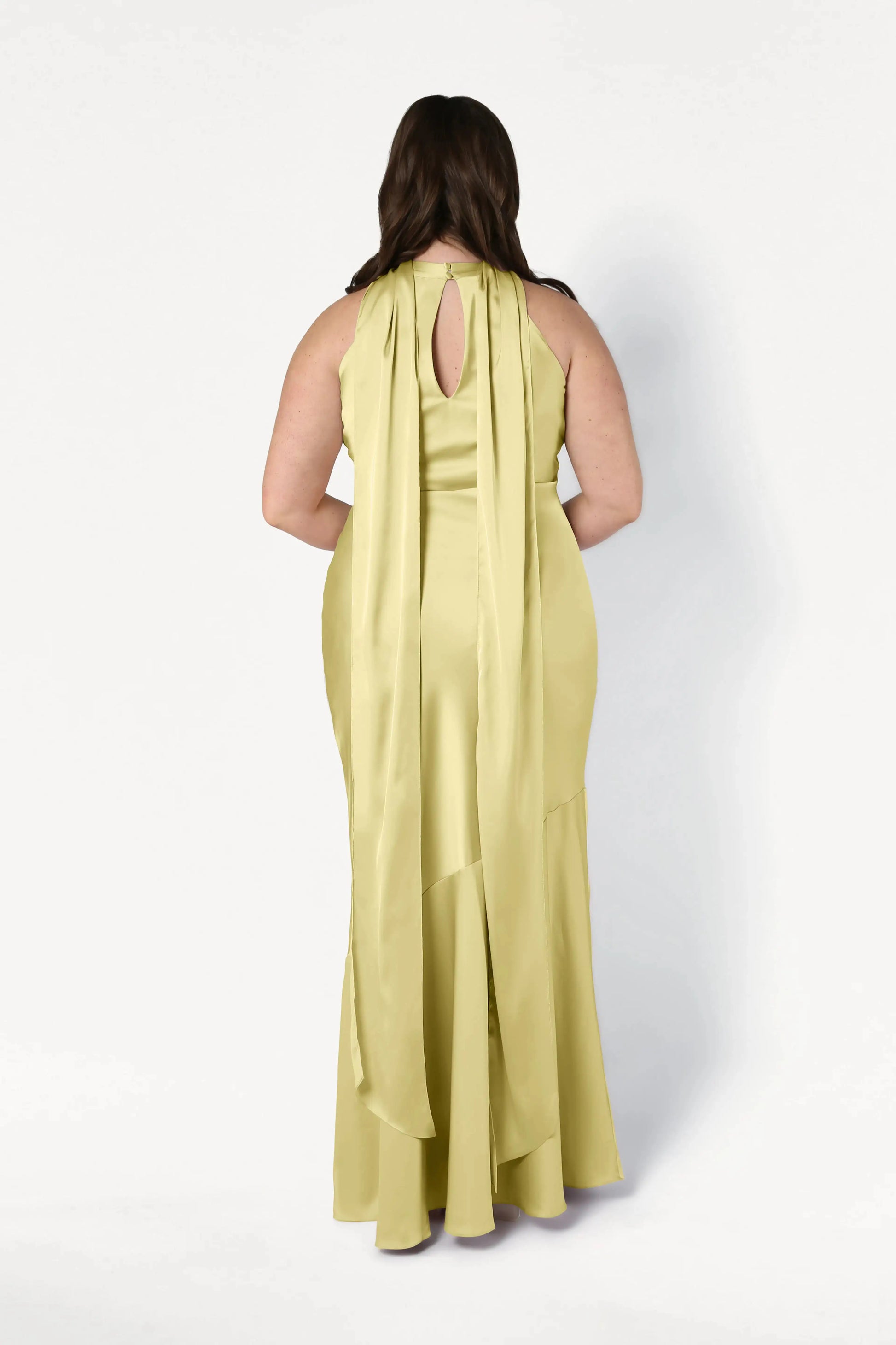A woman stands with her back to the camera, wearing the TH&TH Mira Bridesmaid Dress in Limoncello Satin—a long, sleeveless bias-cut gown with an upper back keyhole and two draped panels flowing to the floor.