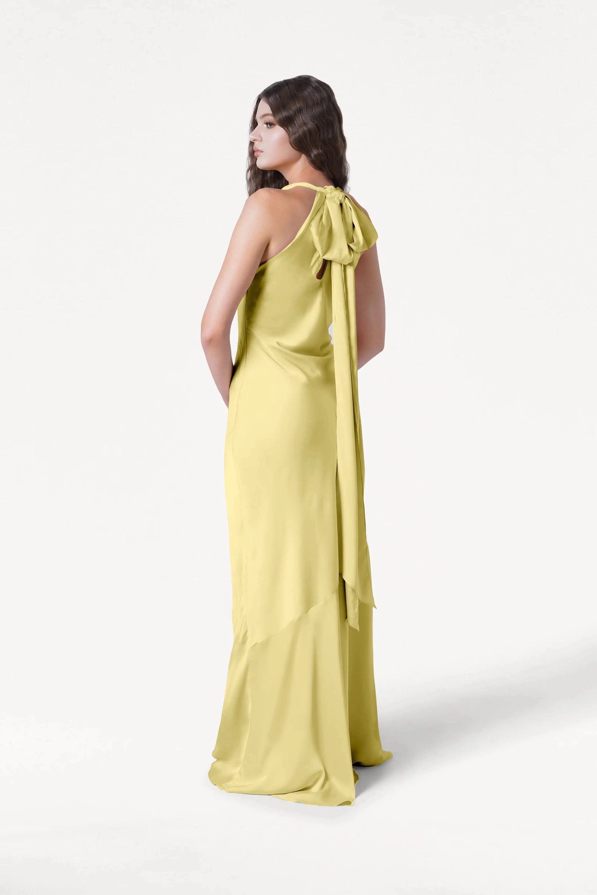 A woman models the TH&TH Mira Bridesmaid Dress in Limoncello Satin, featuring a long silhouette with a large back bow and flowing ribbons, as she stands slightly turned and looks over her shoulder against a white background.