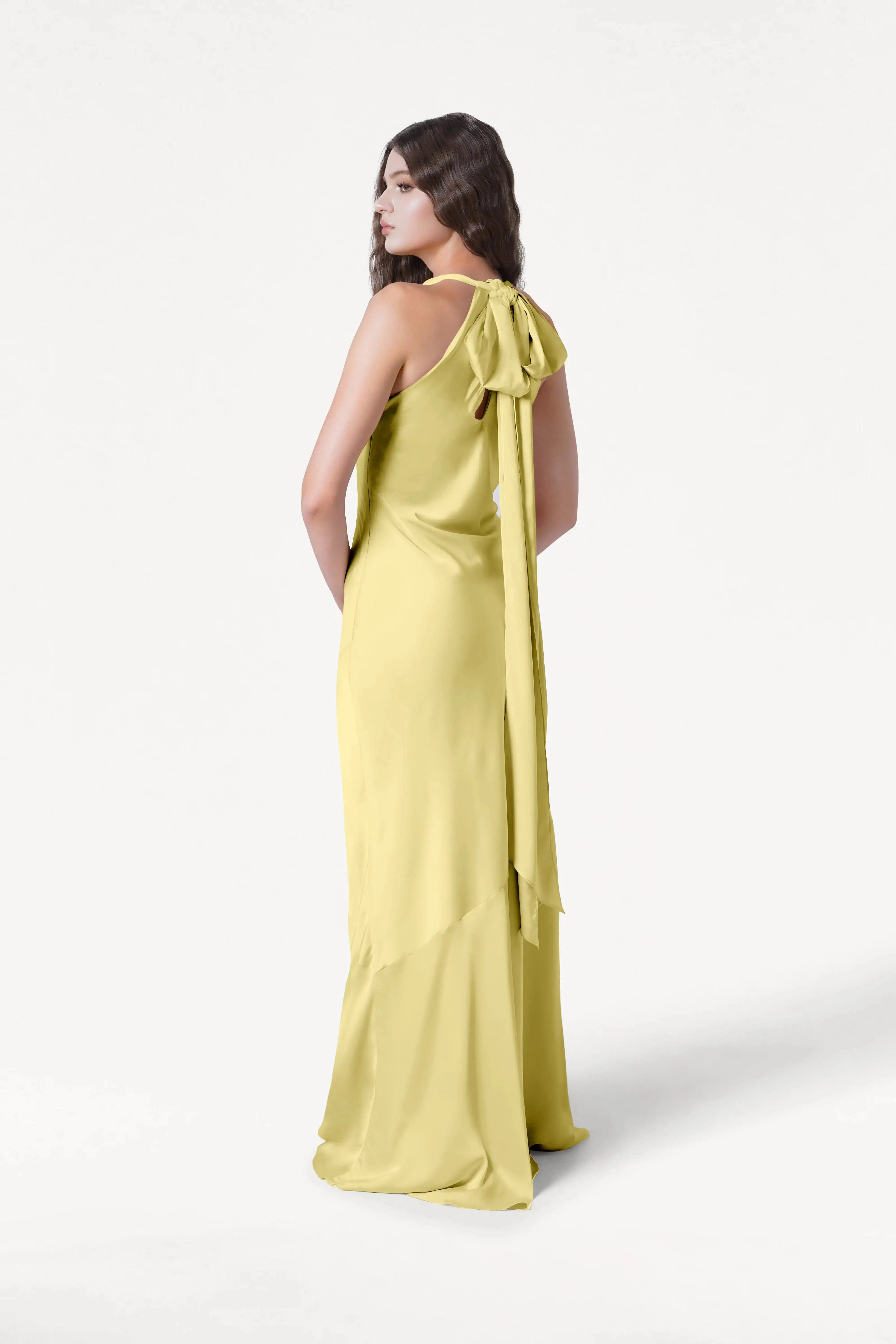 A woman models the TH&TH Mira Bridesmaid Dress in Limoncello Satin, featuring a long silhouette with a large back bow and flowing ribbons, as she stands slightly turned and looks over her shoulder against a white background.
