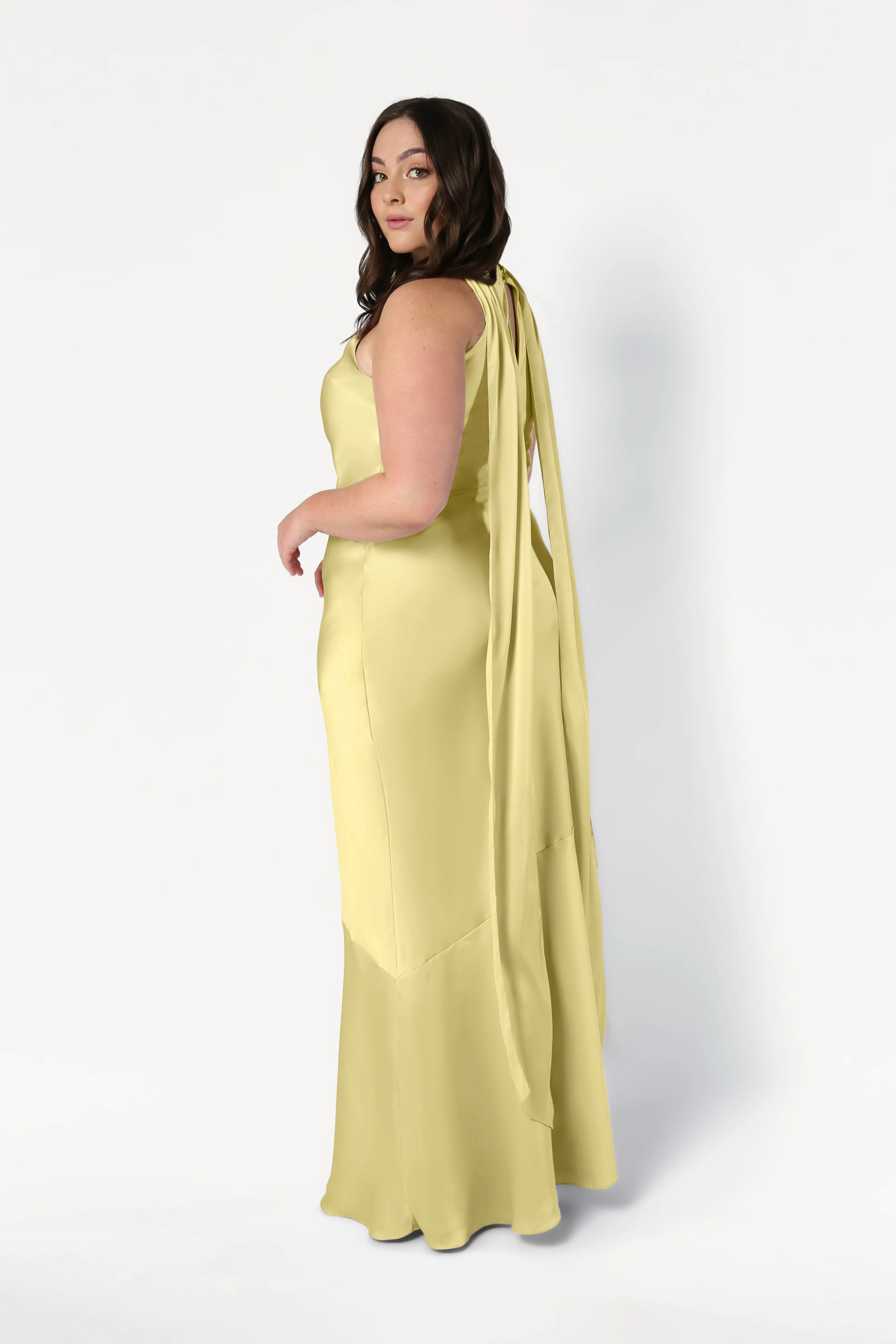 A woman with long brown hair models the TH&TH Mira Bridesmaid Dress in Limoncello Satin—a sleeveless, floor-length gown with a bias-cut silhouette and long sashes at the shoulders—standing against a plain white background.