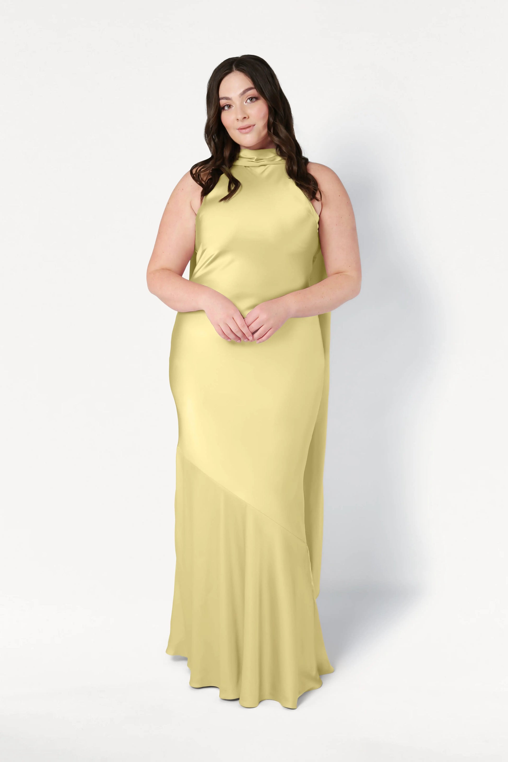 A woman with long dark hair stands against a white background, wearing the TH&TH Mira Bridesmaid Dress in Limoncello Satin—a sleeveless, floor-length halterneck style with a high neckline and subtle diagonal seam detail. Her hands are gently clasped.