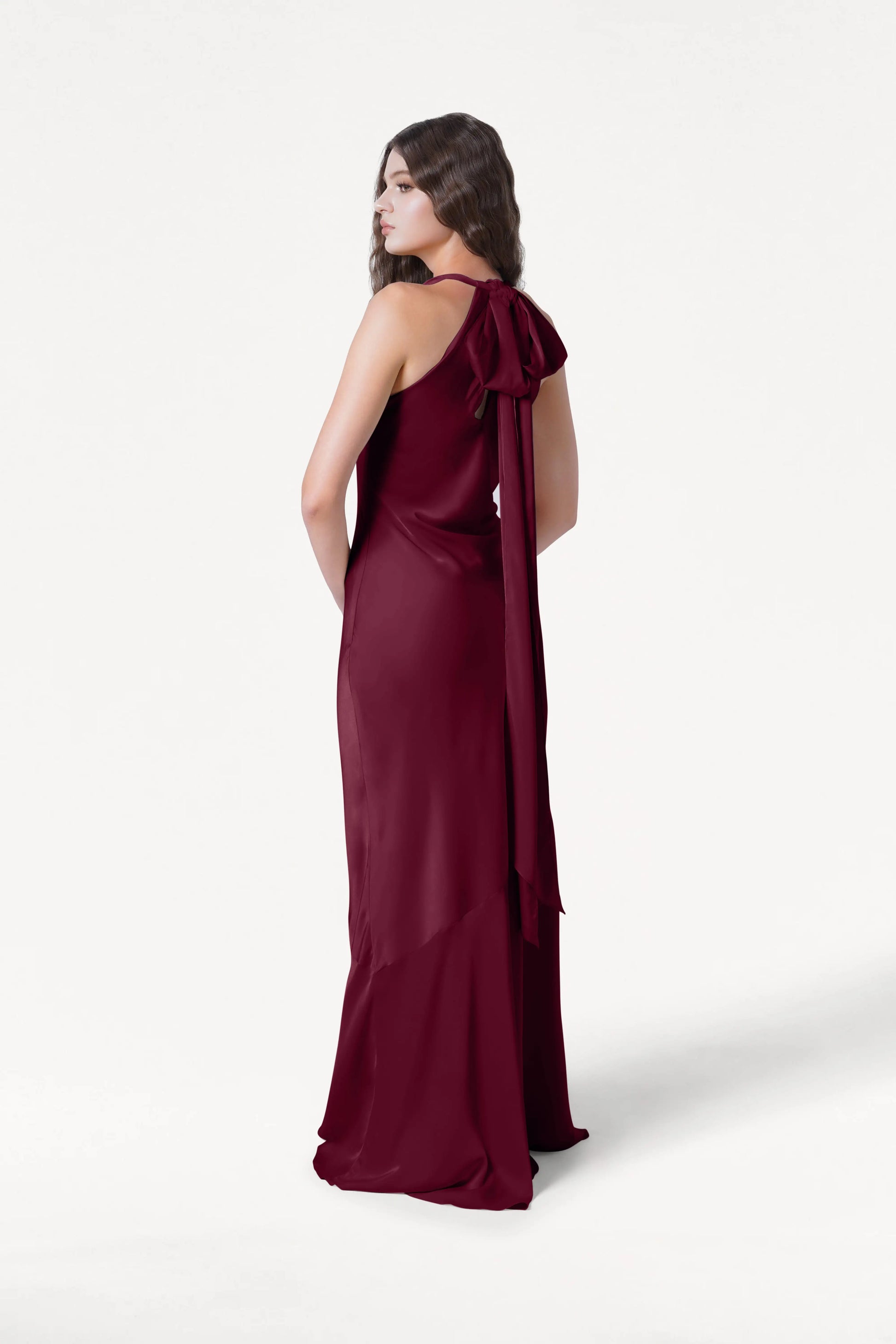 A woman stands against a white background, looking over her shoulder while wearing the TH&TH Mira Bridesmaid Dress in Merlot Satin—sleeveless, floor-length satin with a long bow detail at the back.