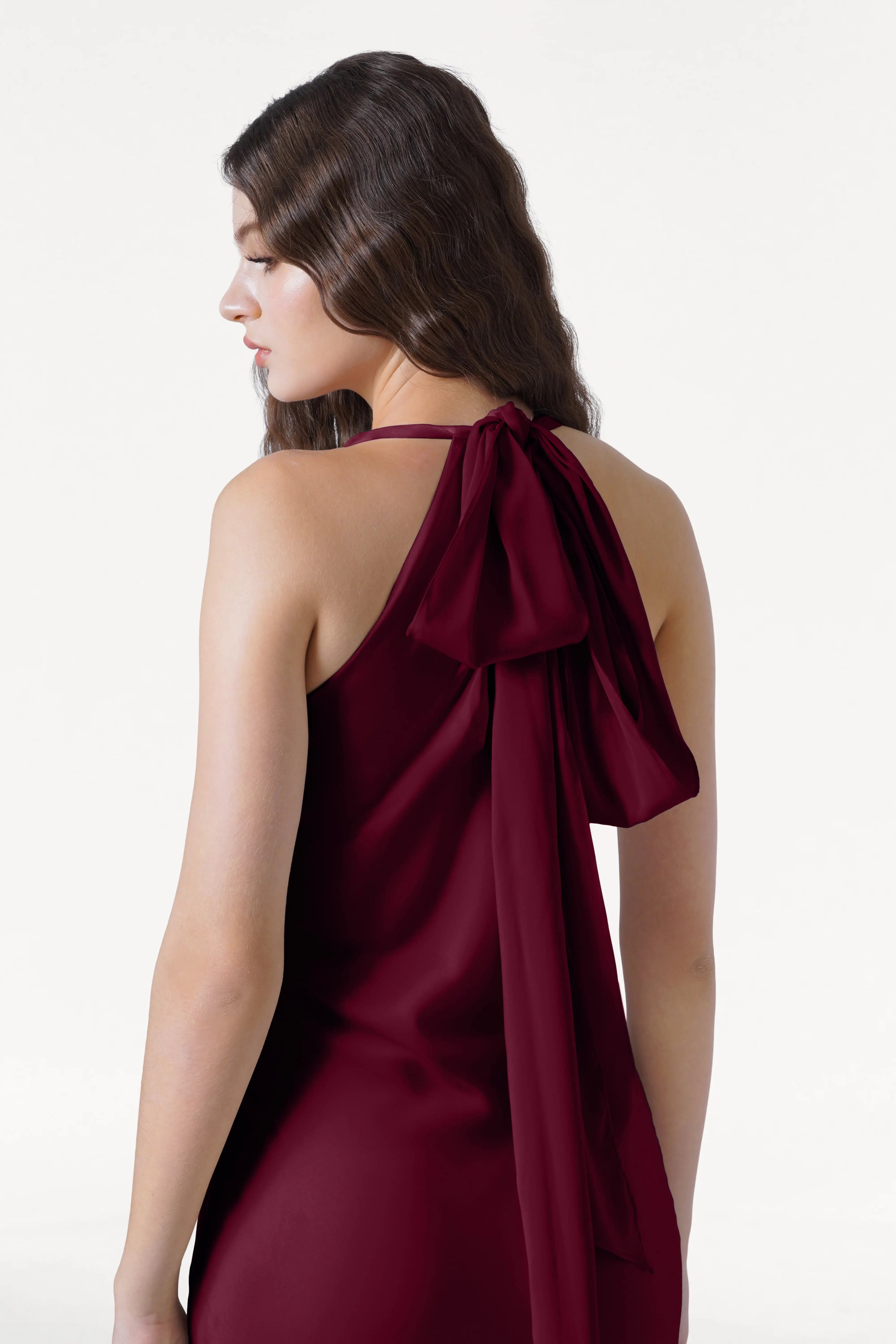 A woman with long, wavy brown hair models the TH&TH Mira Bridesmaid Dress in Merlot Satin. The sleeveless gown features a large back bow and is shown from behind against a plain white background, with her slightly in profile.