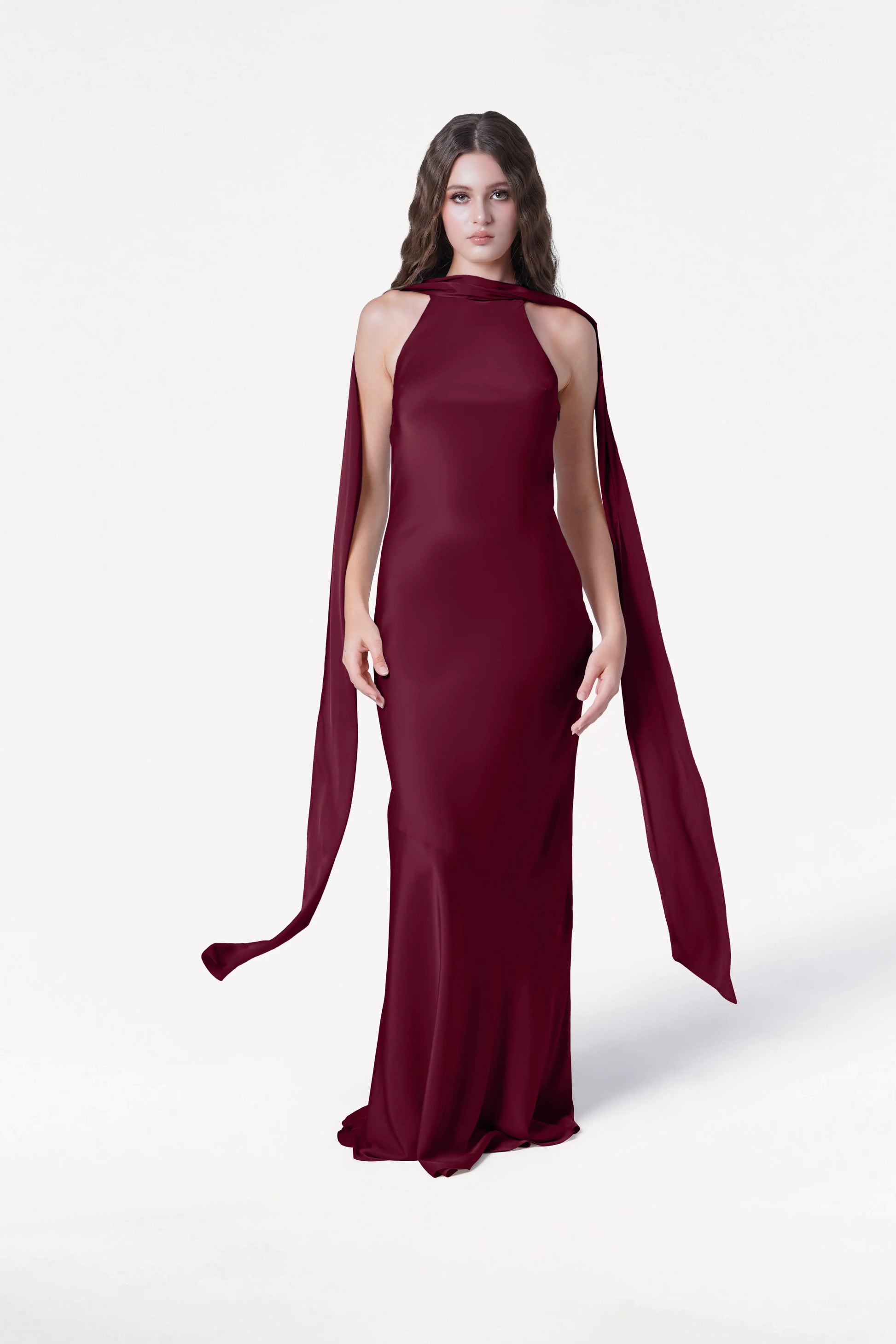 A woman with long, wavy hair stands against a white background wearing the Mira Bridesmaid Dress in Merlot Satin by TH&TH, featuring a fitted silhouette and cape-style draping from her shoulders as she looks directly at the camera.