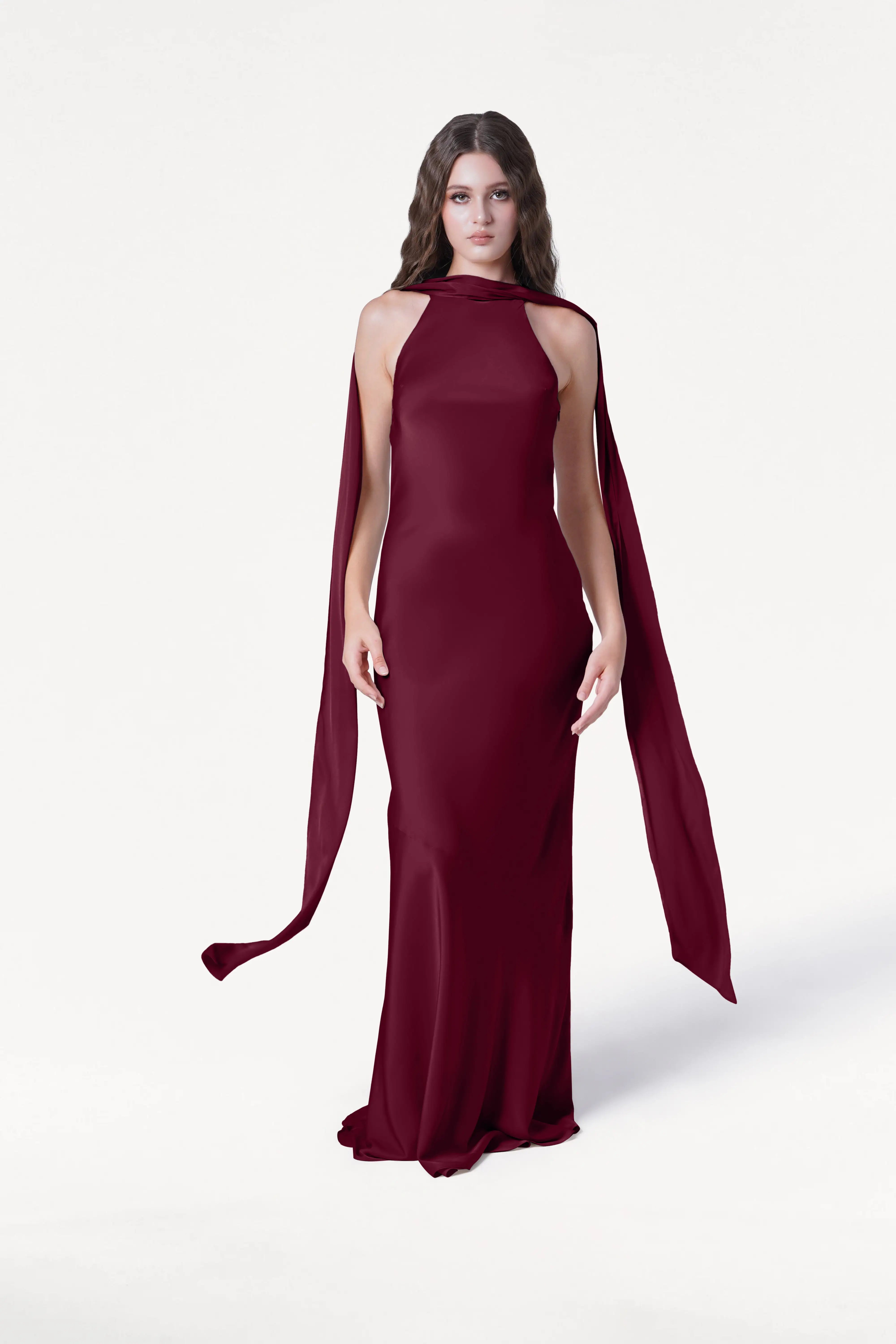 A woman with long, wavy hair stands against a white background wearing the Mira Bridesmaid Dress in Merlot Satin by TH&TH, featuring a fitted silhouette and cape-style draping from her shoulders as she looks directly at the camera.