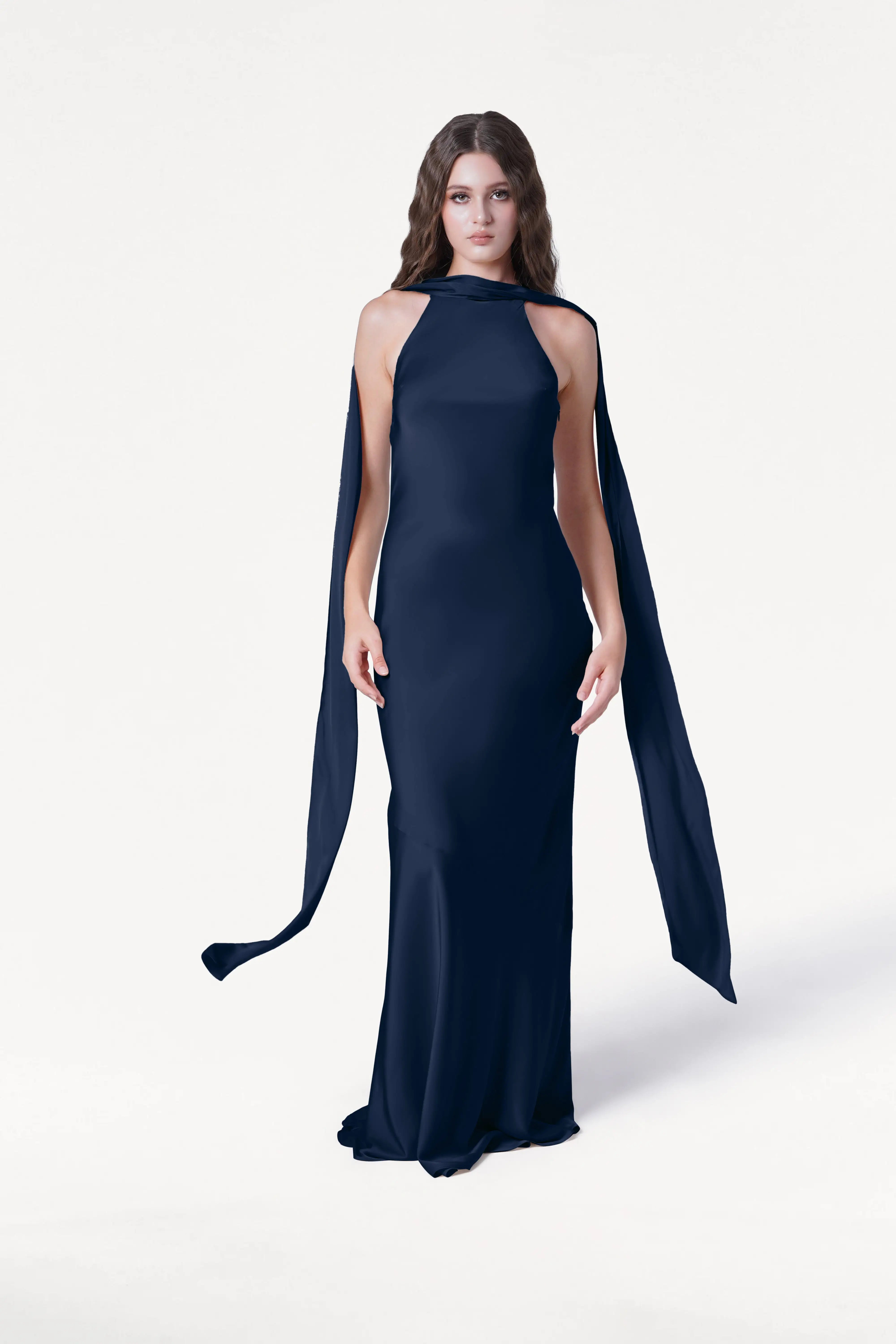 A woman with long brown hair wears the TH&TH Mira Bridesmaid Dress in navy satin, featuring a sleeveless floor-length silhouette, high neckline, and flowing cape-like panels, standing against a plain white background.