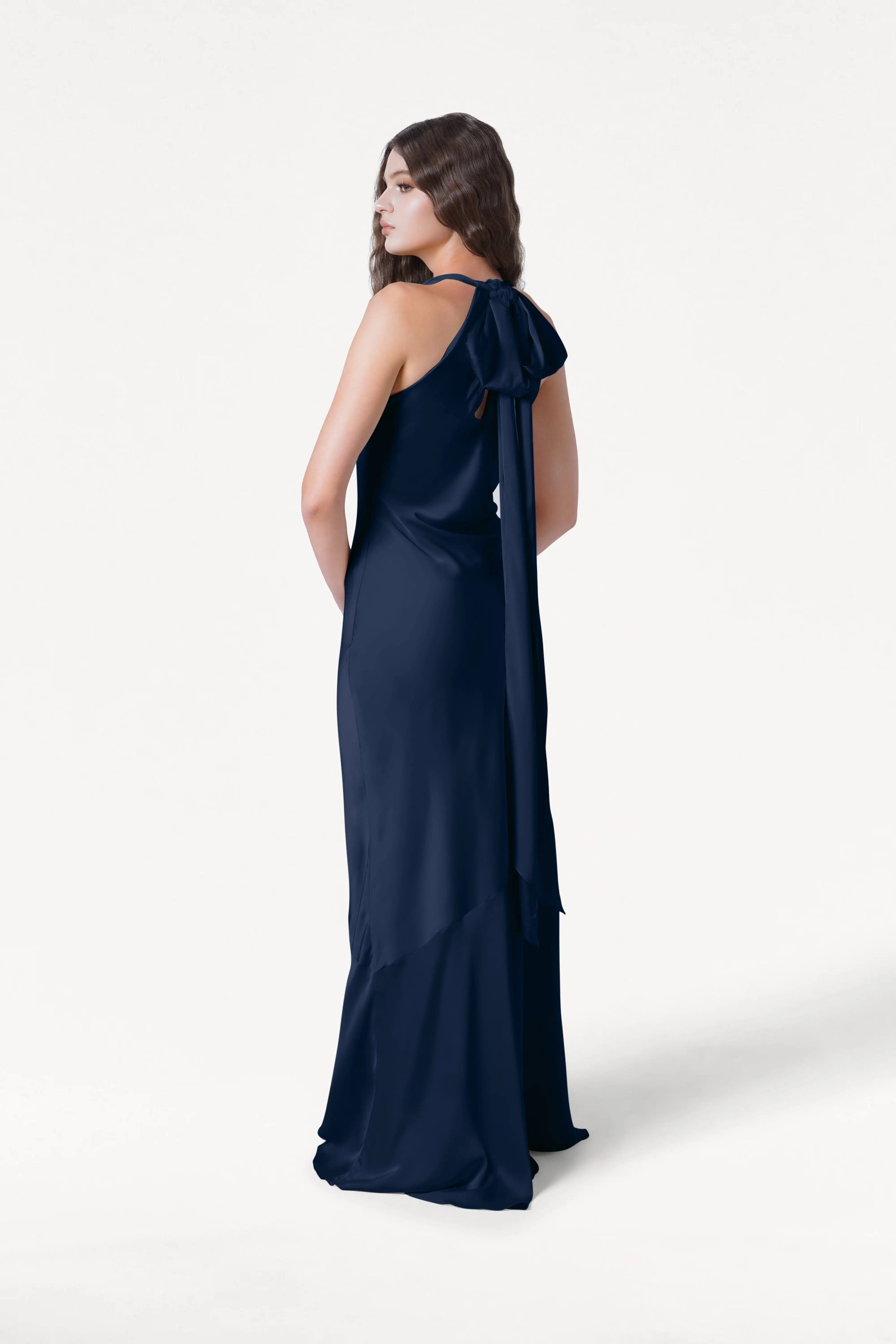 A woman faces away in the Mira Bridesmaid Dress in Navy Satin by TH&TH, a bias-cut gown with a flowing ribbon tied at the nape, set against a plain white background.