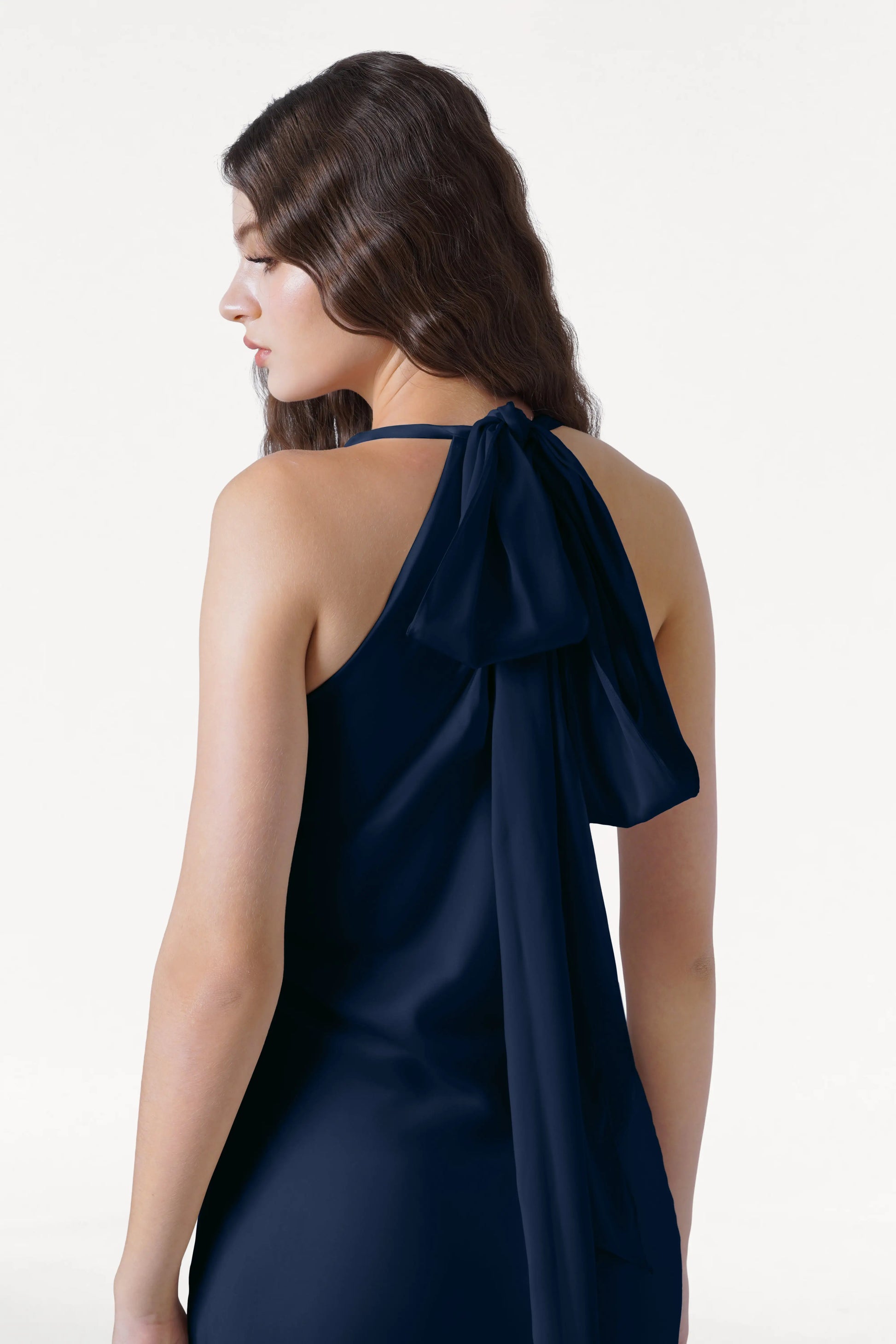 A woman with long wavy brown hair is seen from behind wearing the TH&TH Mira Bridesmaid Dress in navy satin, a sleeveless halterneck style with a large bow at the neckline, set against a plain white background.