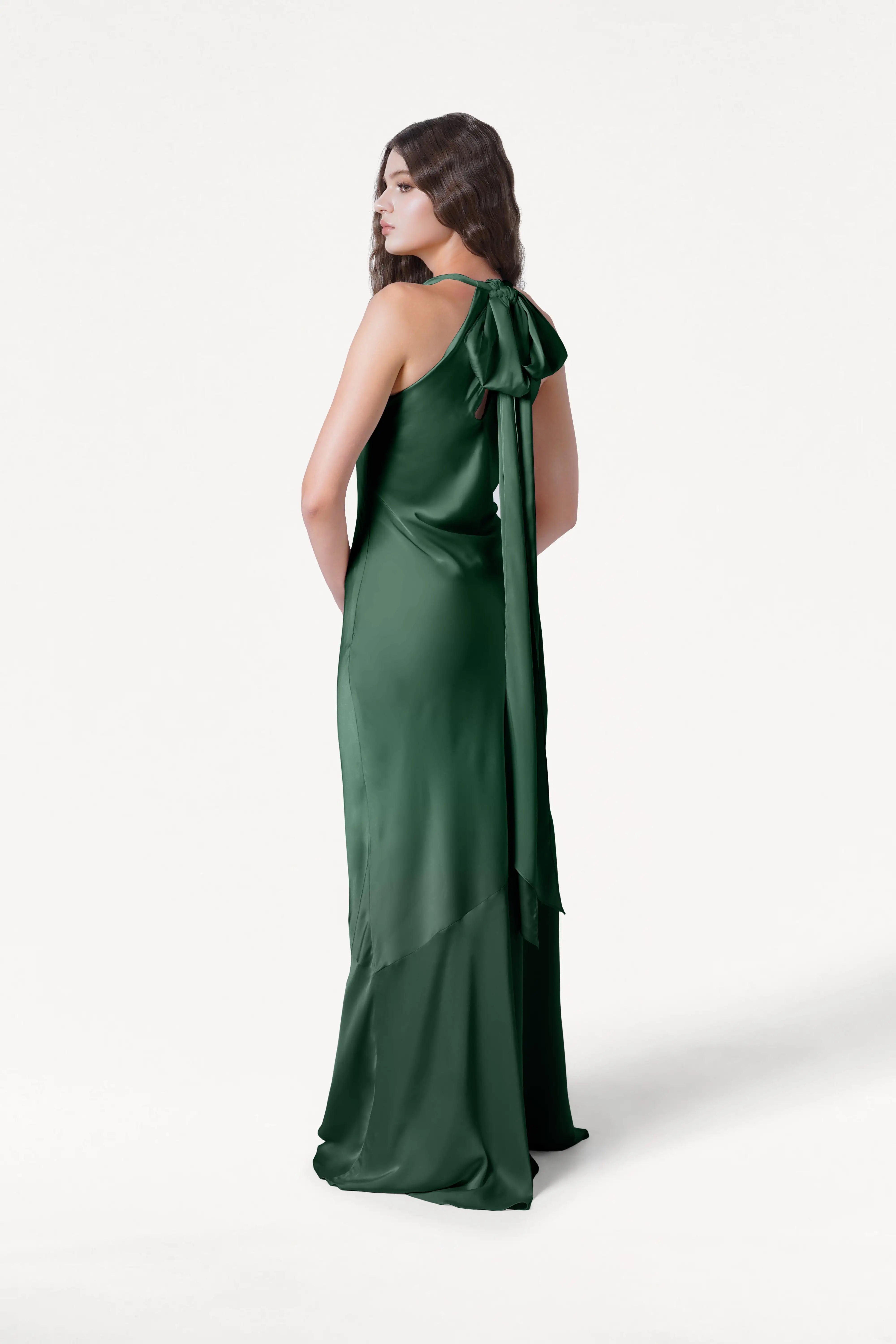 A woman stands against a white background, looking over her shoulder in the Mira Bridesmaid Dress in Olive Grove Satin by TH&TH—a long, sleeveless gown featuring a large back bow.
