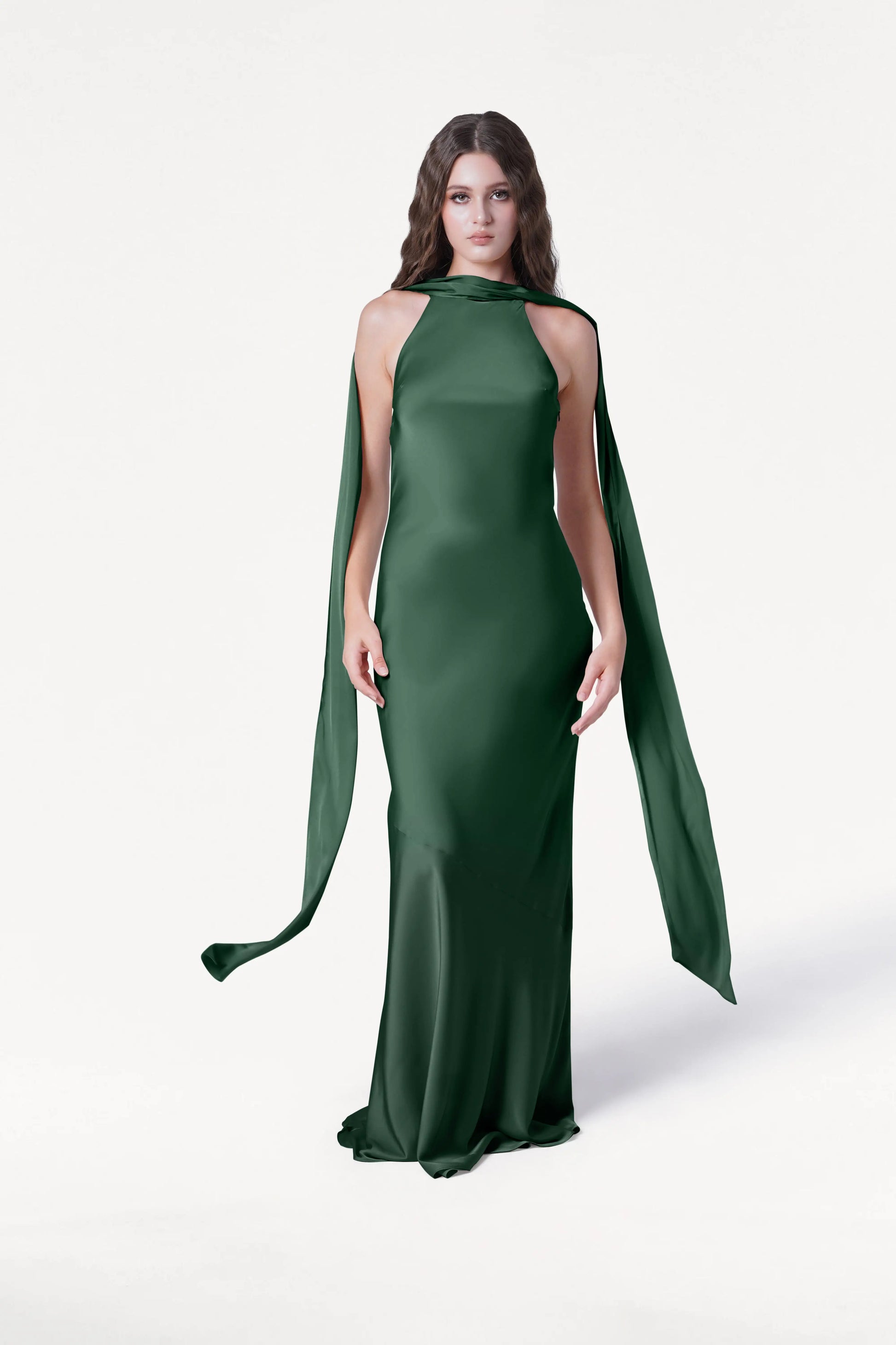 A woman with long brown hair and a neutral expression wears the TH&TH Mira Bridesmaid Dress in Olive Grove Satin, featuring a floor-length silhouette and elegant, scarf-like extensions, against a plain white background.