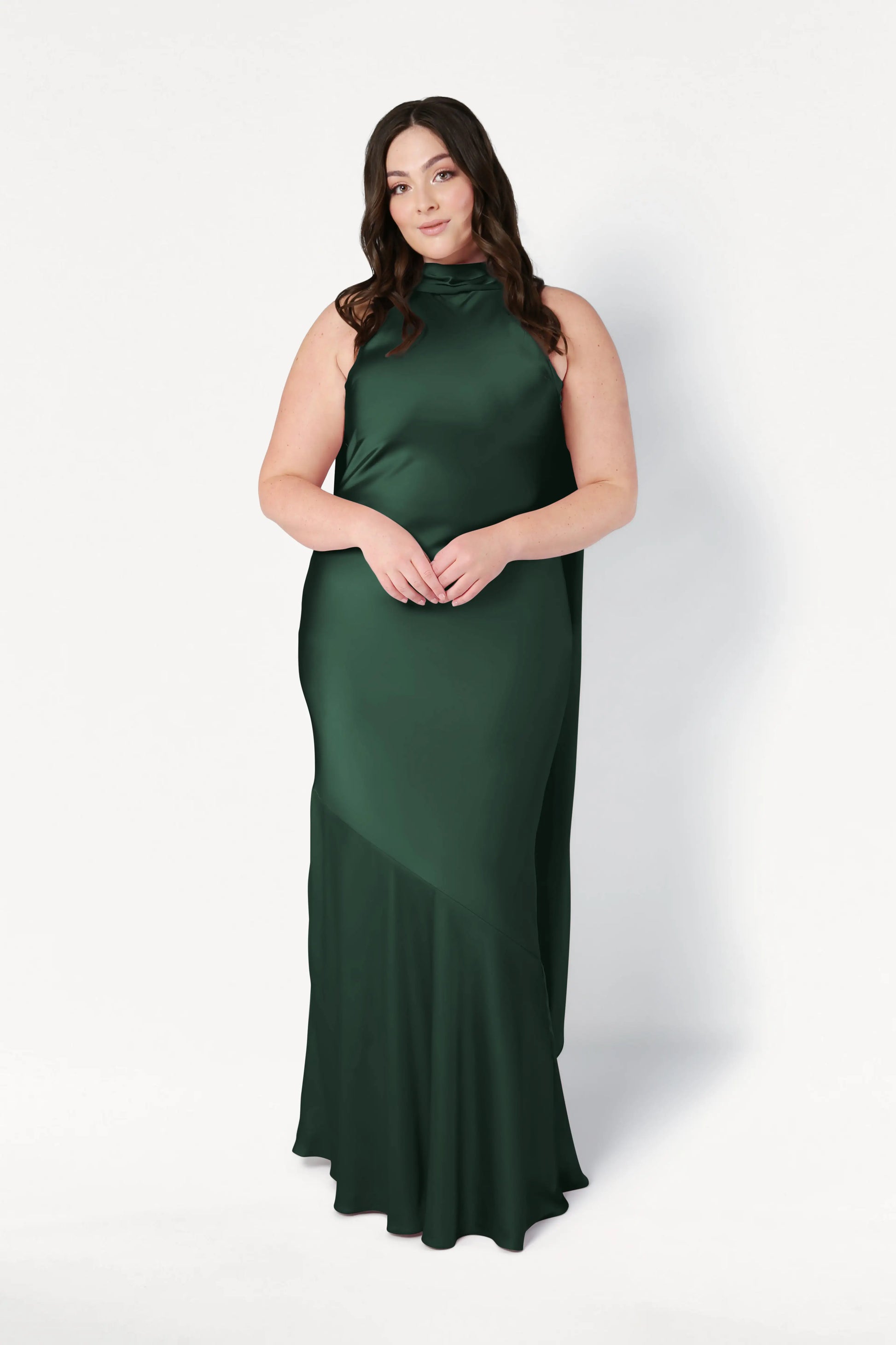 A woman with long brown hair in loose waves wears the TH&TH Mira Bridesmaid Dress in Olive Grove Satin, a sleeveless, floor-length gown with a high neckline, standing against a plain white background.