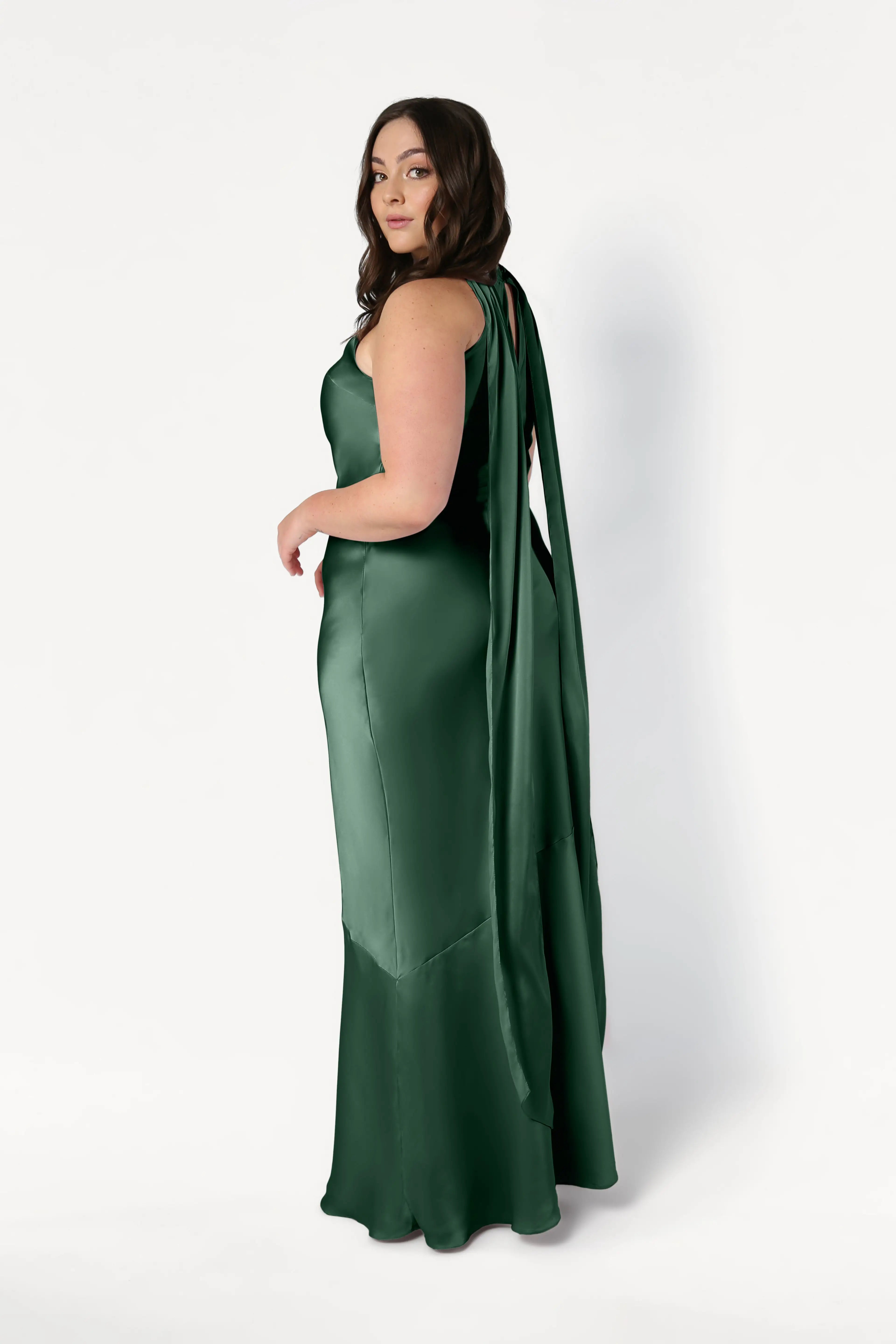 A woman with long brown hair wears the TH&TH Mira Bridesmaid Dress in Olive Grove Satin, featuring a sleeveless, floor-length silhouette and draped back detail, standing against a plain white background and looking over her shoulder.