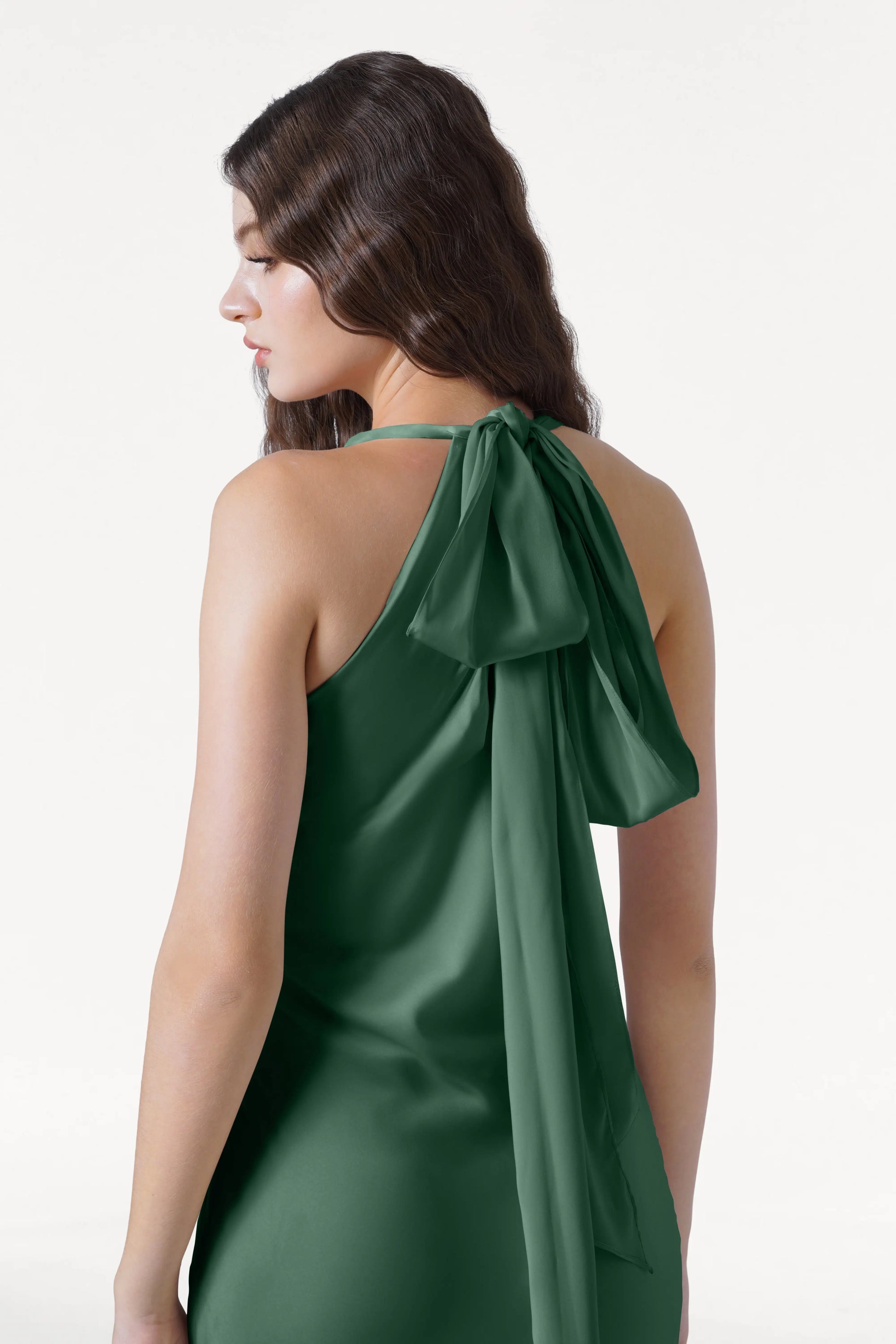 A woman with long, wavy brown hair models the TH&TH Mira Bridesmaid Dress in Olive Grove Satin, featuring a halterneck and large neck bow, displaying its smooth satin texture against a white background.