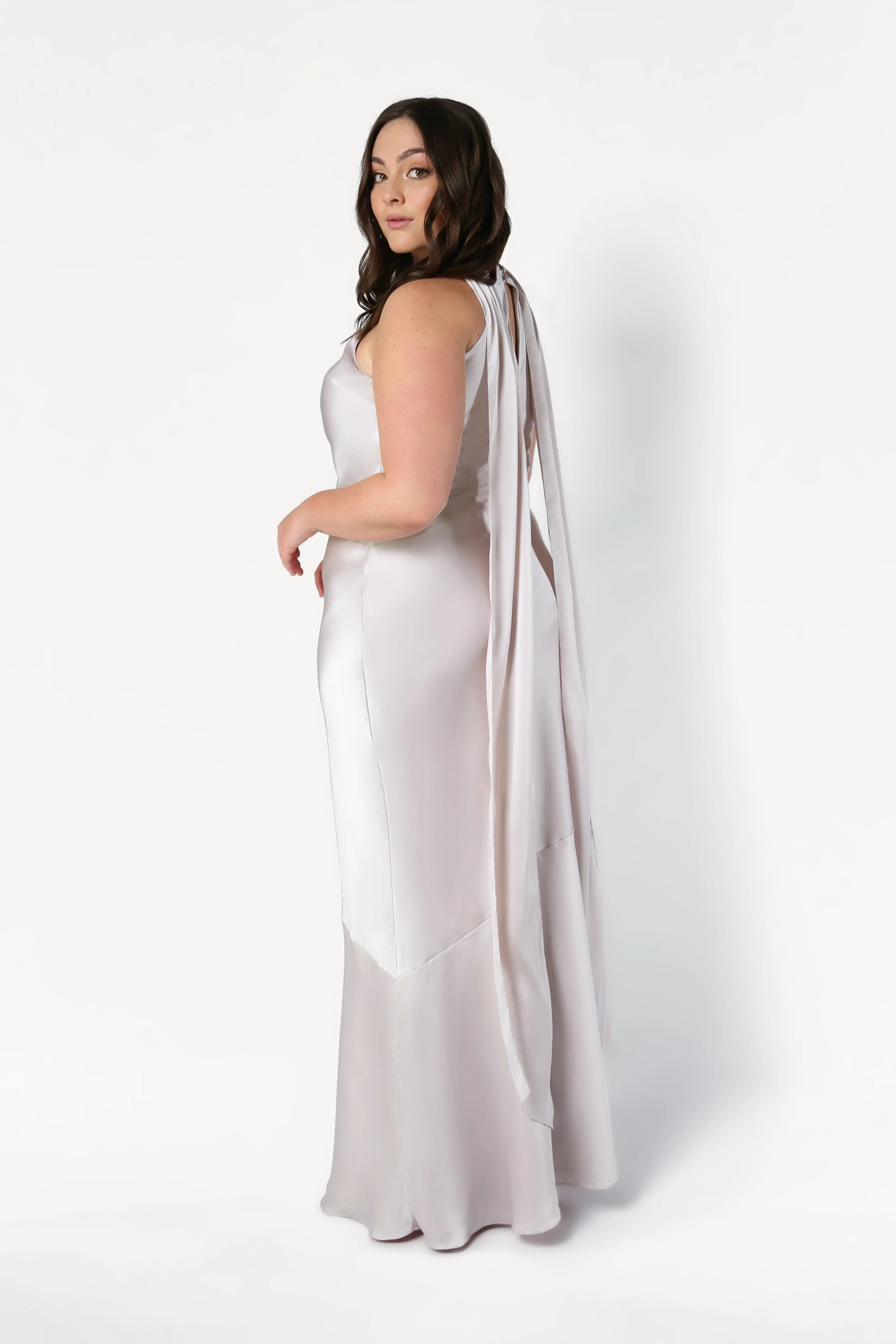 A woman with wavy brown hair models the Mira Bridesmaid Dress in Oyster Satin by TH&TH, featuring a long, elegant silhouette with flowing back detail. She stands sideways and looks over her shoulder against a white background.