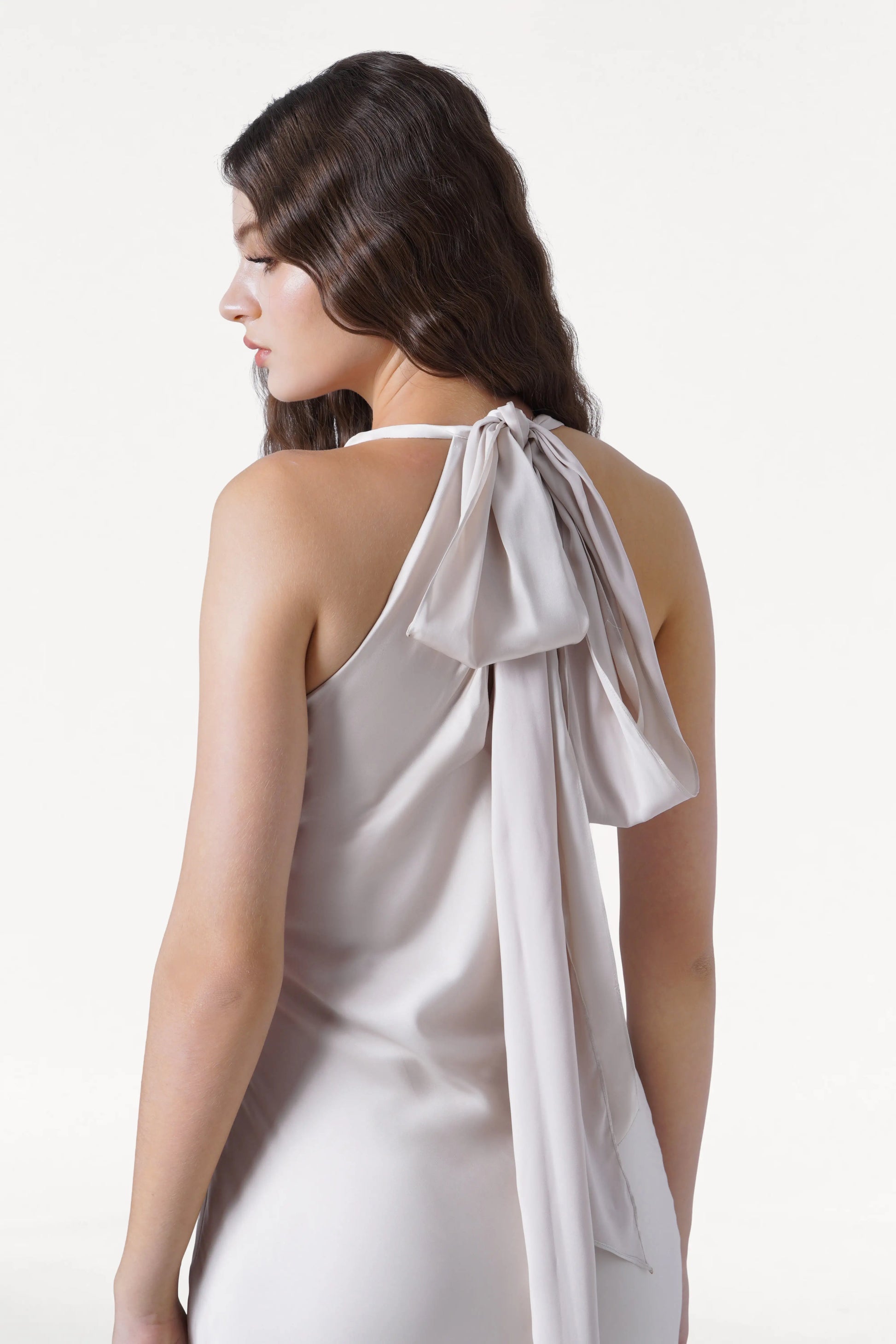 A woman with long, wavy brown hair is seen from the side and back wearing the TH&TH Mira Bridesmaid Dress in Oyster Satin, featuring a bias cut and a large bow tied at the back of her neck. The background is plain white.